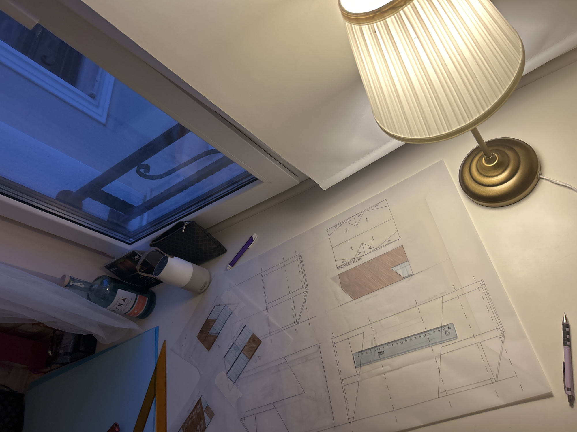 The image displays a desk next to a window, illuminated by a desk lamp. Architectural blueprints are spread across the desk, with a ruler and pens for detailed work. In the background, a bottle of "SYKA" vodka and a small portable speaker are visible. The window offers a glimpse of a balcony railing and the dimly lit exterior. The overall impression is of a workspace for design or architecture.