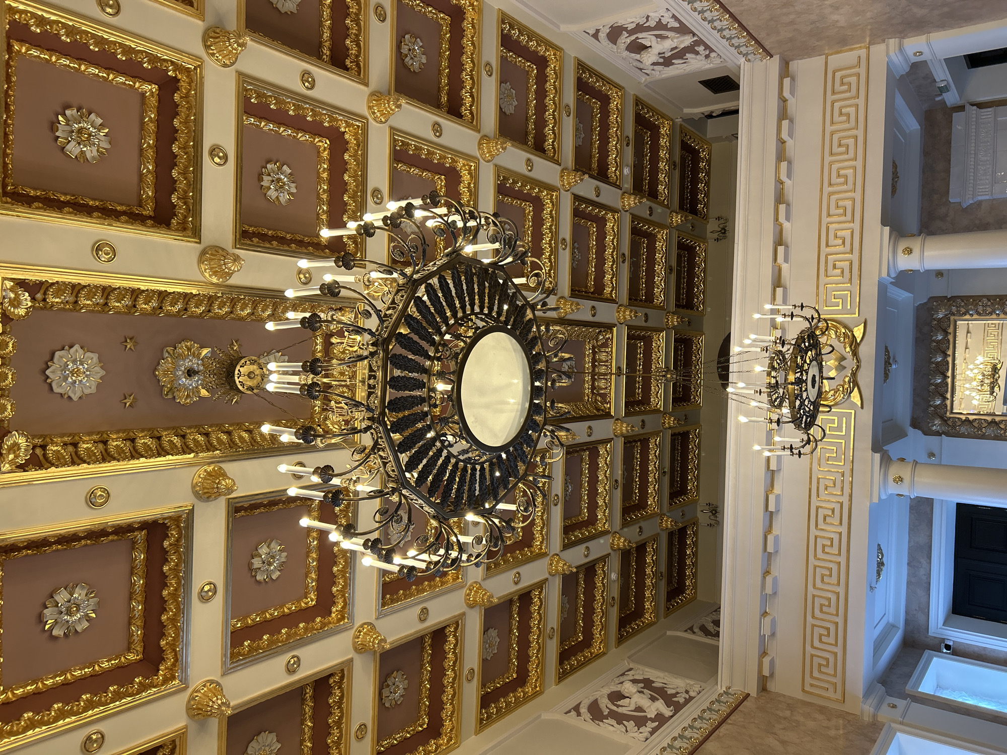 The image captures the ornate interior of a grand building, likely a palace or historical hall. The ceiling is a dominant feature, heavily decorated with intricate gilded molding, creating a coffered effect. Each coffer is outlined in gold and features a central decorative element, some appearing to be floral motifs or rosettes with white and silver accents. The ceiling also has a reddish-brown hue in some sections, contrasting with the gold and cream colors.
Two large chandeliers hang from the ceiling, both appearing to be made of dark metal and adorned with numerous candle-like lights. The upper chandelier is larger and more elaborate, with multiple tiers and a central circular light fixture. The lower chandelier is simpler, with a circular arrangement of lights.
Below the main ceiling, a mezzanine or balcony level is visible, separated by a white balustrade with a gold Greek key pattern. White columns support the upper structure. The walls appear to have a textured finish in muted tones, and there are some white sculptural reliefs on the upper walls, depicting figures that appear to be cherubs or mythological beings. A mirror with a gilded frame is also visible on the wall below the mezzanine. The overall impression is one of opulence and historical grandeur.
The provided metadata states the location is in "Добасненскі сельскі Савет, Belarus." Based on the visual evidence, this scene is an indoor architectural space characterized by elaborate classical or baroque decorative elements. There are no people visible in the image.