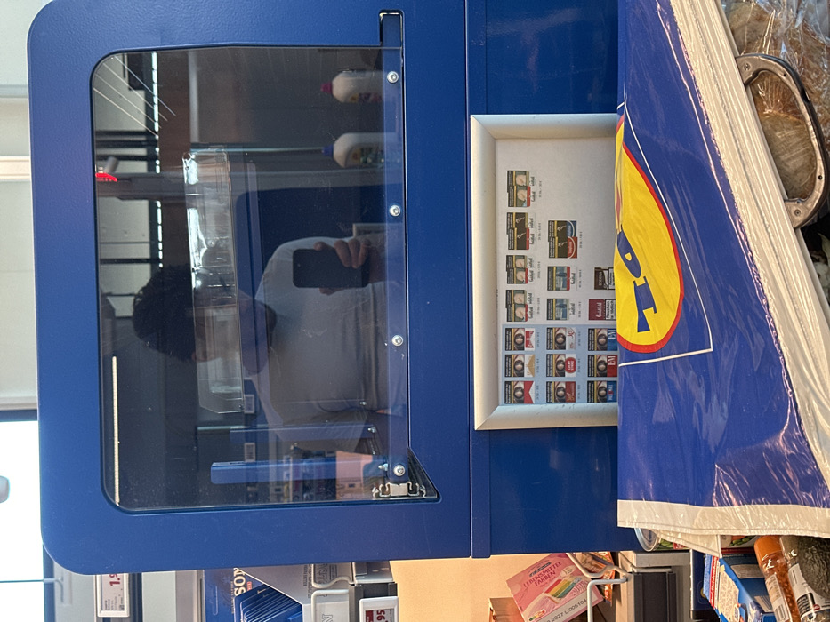 The scene depicts the checkout area inside a Lidl supermarket located in Jestetten, Germany. The viewpoint is from a first-person perspective, looking towards the cashier's station.
In the foreground, a stack of blue plastic shopping bags with the yellow "Lidl" logo is prominently displayed, partially obscuring groceries such as an avocado and other packaged items on what appears to be a conveyor belt or bagging area.
Above the shopping bags, a blue structure, likely part of the checkout counter, features a clear, transparent protective screen secured with multiple metal fasteners. Reflected within this screen is the upper torso and head of a person with dark, curly hair, wearing a light-colored t-shirt. This individual is holding a black smartphone with their right hand, indicating they are taking the photograph. The reflection also shows parts of the supermarket interior, including fluorescent lighting fixtures and two white plastic bottles with colored caps.
Below the protective screen, integrated into the blue counter structure, is a framed display showcasing various tobacco products, including cigarettes from brands such as Marlboro, Lucky Strike, Goldfield, Roth-Händle, EM, HB, and Bodega. Each product image includes German text warning labels and pricing (e.g., "20 St. / 7,20 €"). To the left of this display, a rack containing rolling papers, with a "SONY" branded package partially visible, displays price tags in Euros (e.g., "1,99" and "1,95"). A pink item, possibly a flyer or magazine, is also present on the far left.