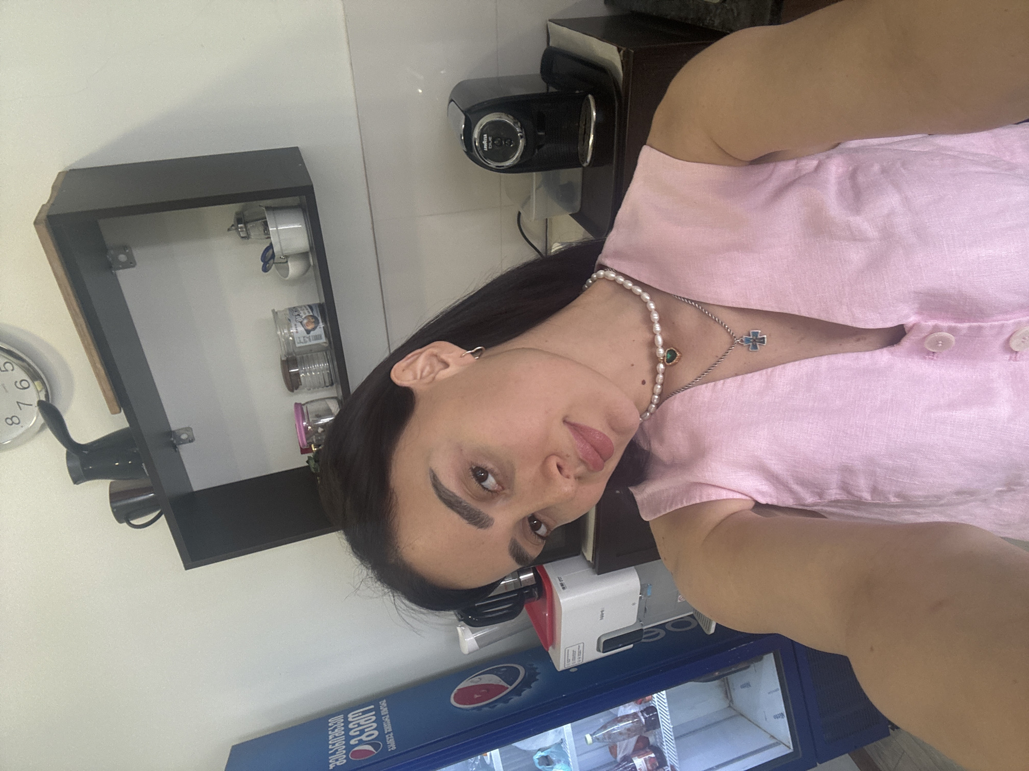 A person is visible in the frame, wearing a pink top, a pearl necklace, and a silver cross necklace. They are positioned in front of a blue beverage cooler with "Coca Cola" branding written in Georgian. Beside the cooler is a white appliance. In the background, a dark-colored shelf unit hangs on the wall, displaying cups and jars. A black espresso machine is also visible on a dark countertop. The setting appears to be indoors, possibly a kitchen or small store. The location is inferred to be Tbilisi, Georgia based on the Georgian writing on the Coca-Cola cooler.