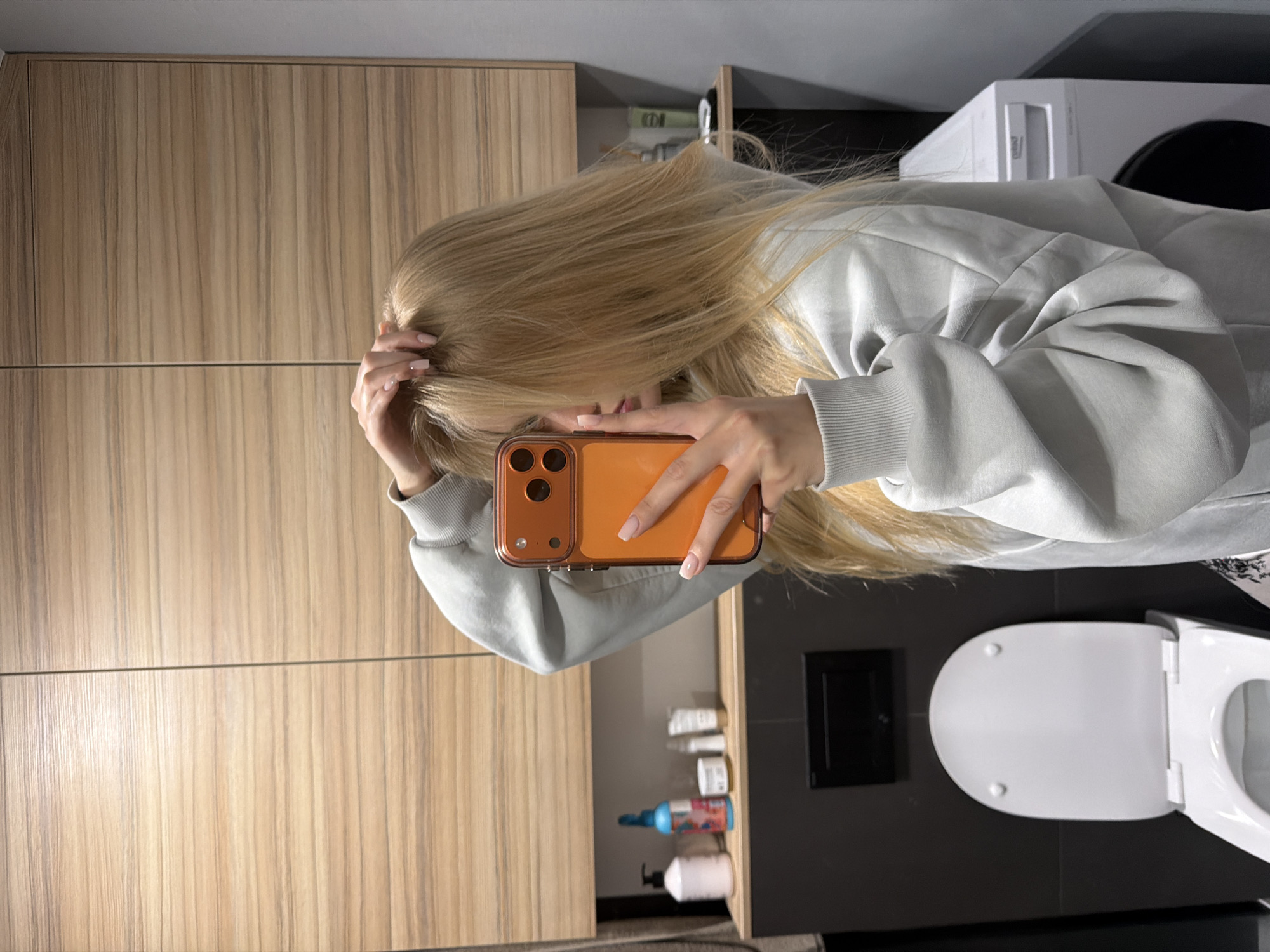A person with long, blonde hair is taking a selfie in what appears to be a bathroom. The person is wearing a light gray sweatshirt. The phone has a clear orange case. The bathroom features light wood-grained cabinets, a white toilet with the lid up, a washing machine, and a black wall with a small square inset. Several bottles of toiletries are visible on a shelf. The location is Unknown, Unknown.