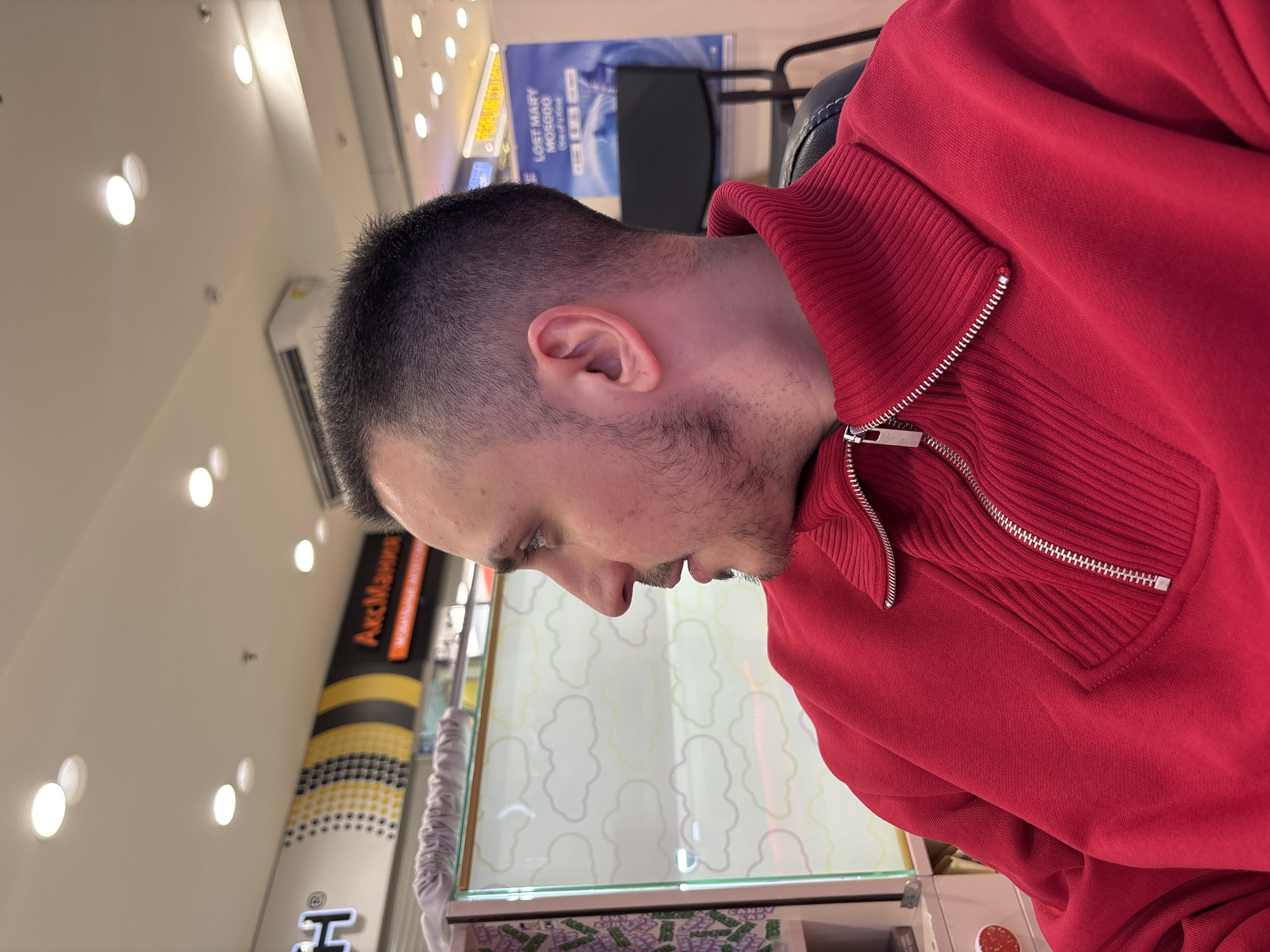 A medium shot depicts a man in a red zip-up sweater, leaning over what appears to be a display case. The display case contains items packaged in purple and white wrappers. A tall, promotional advertisement is visible in the background. The environment is brightly lit with overhead lights. Behind the man, a black office chair and a poster bearing the words "LOST MARY MO5000" are partially visible. Without further information, it is not possible to ascertain if this image was taken in Saint Petersburg, Russia.