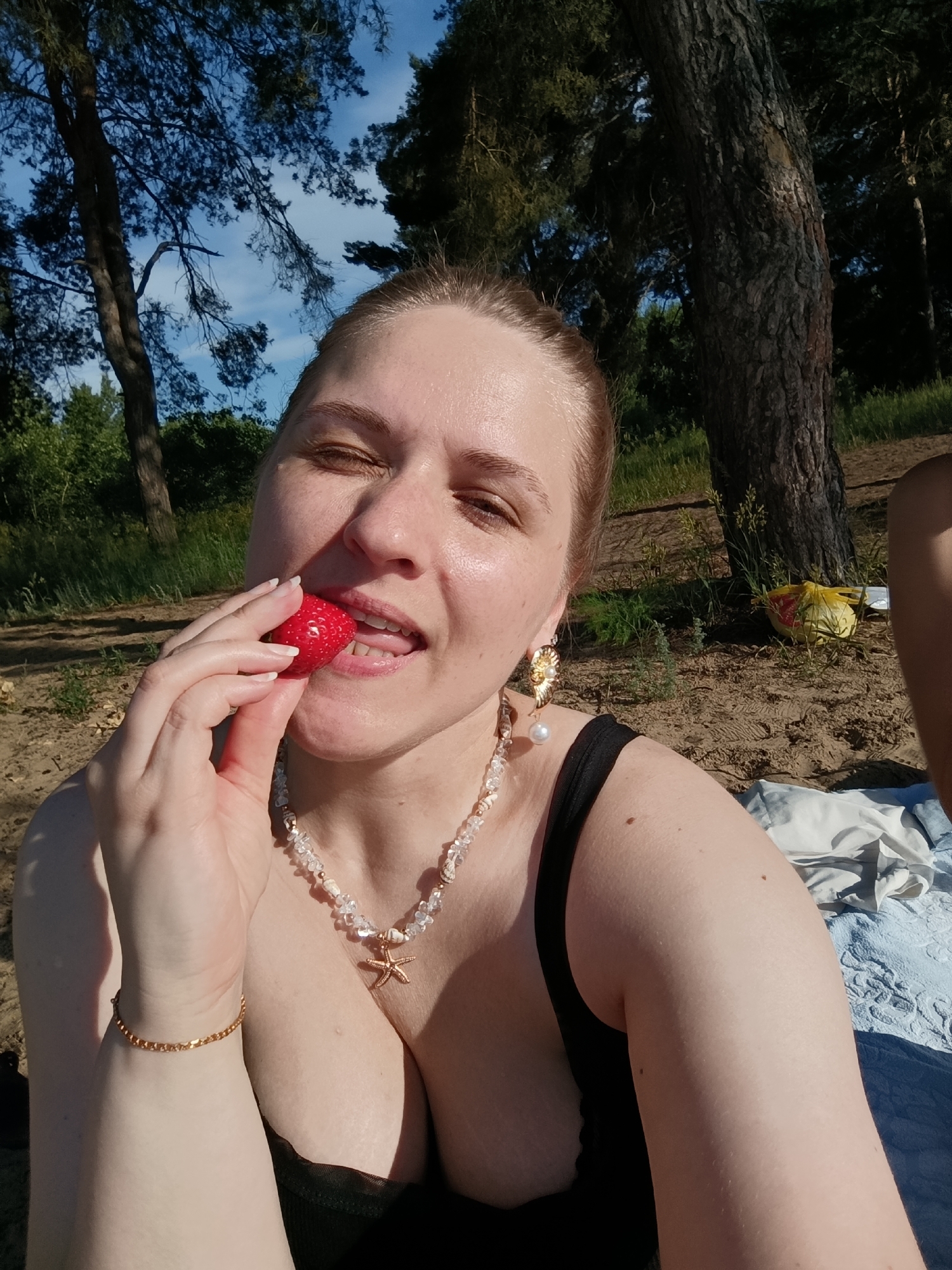 A woman with light skin and blonde hair pulled back is sitting on a sandy embankment in a park or forest setting, with trees and a blue sky visible in the background. She is wearing a black top and a necklace with a starfish pendant. In her right hand, she holds a strawberry up to her mouth, with her tongue extended. Her eyes are closed. A yellow plastic bag is visible in the background. The location is identified as the city of Подстёпки, Russia.