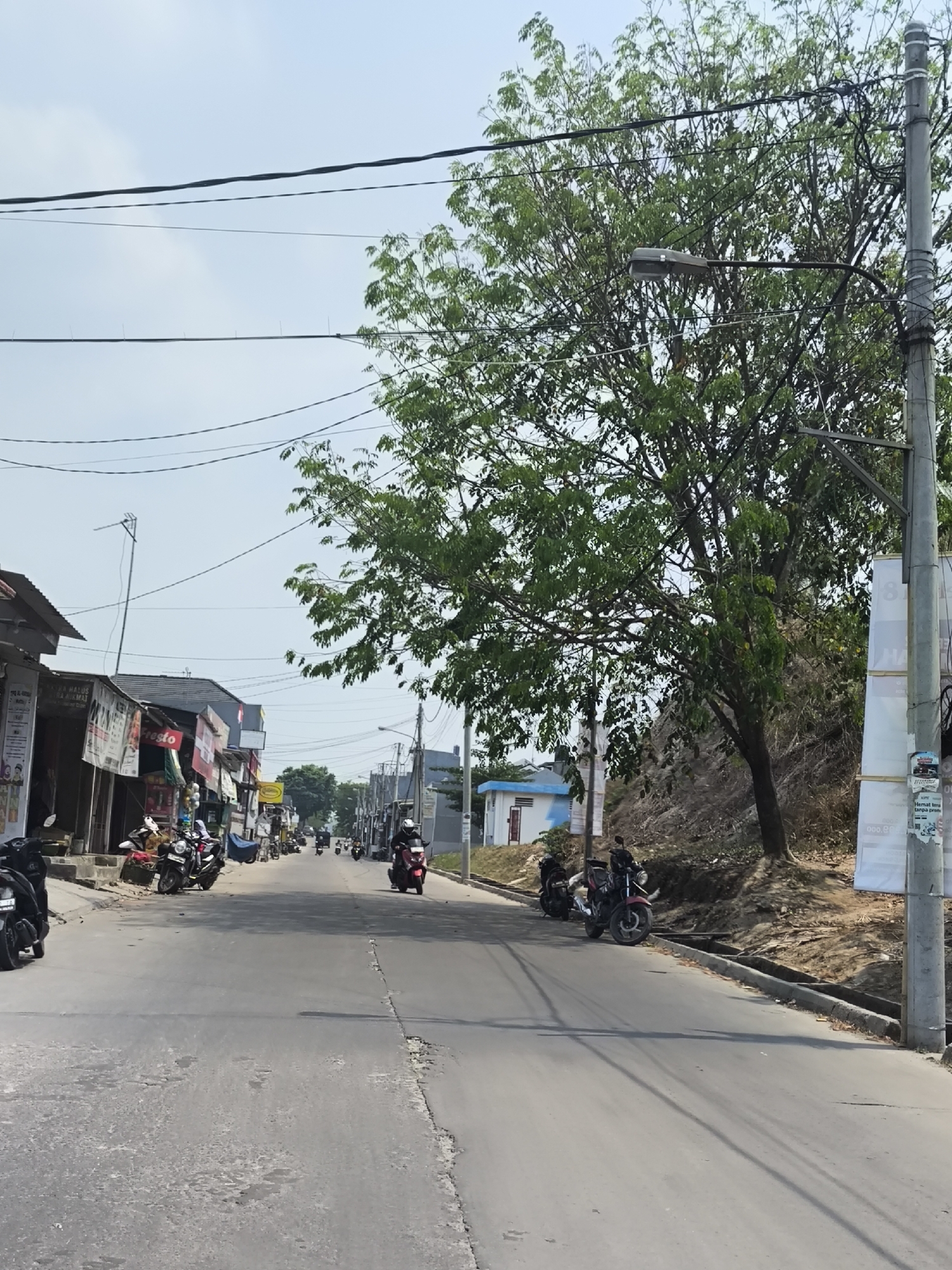 The image depicts a street scene in Cilegon, Indonesia, on a bright, clear day. The road is paved and appears to be in moderate condition with some cracks and patches. On the left side of the road, there are several small shops with their fronts open, displaying various goods and signage. Several motorcycles are parked along the sidewalk in front of these shops.
On the right side of the road, a large tree with lush green foliage dominates the mid-ground. Beyond the tree, a slope covered with dry vegetation and dirt can be seen. A concrete utility pole with overhead wires stands prominently in the foreground on the right, with posters attached to it. Further down the street, more buildings and shops are visible, along with a few more motorcycles and what appear to be individuals riding them, suggesting moderate traffic. In the distance, the sky is clear and light blue. The overall impression is of a typical urban or semi-urban street environment in Indonesia.
