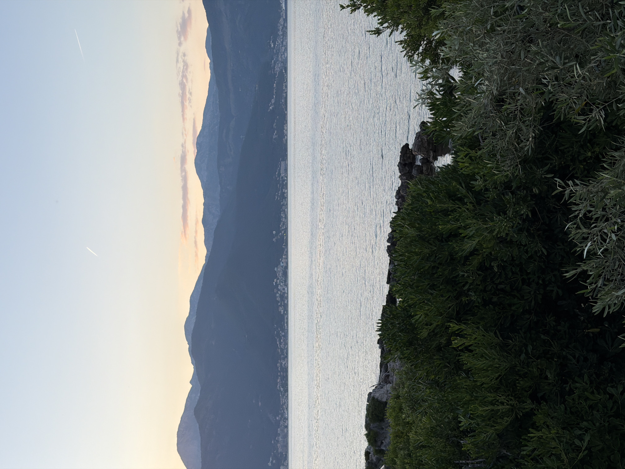 The image shows a body of water with mountains in the background. The location appears to be the city of Gošići, Montenegro, based on the geographical features. On the right of the body of water, there is lush greenery. The sky is a pale color with a white streak. The mountains in the background are gray.
