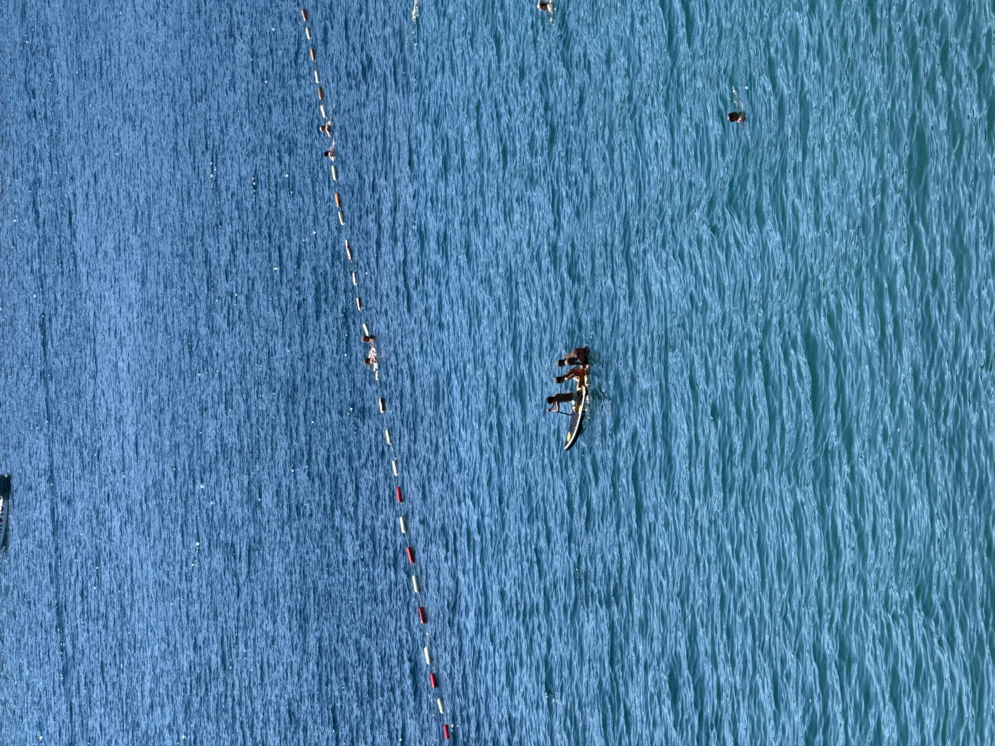 The image depicts a wide expanse of deep blue, rippling ocean water, likely off the coast of Budva, Montenegro, under bright daylight. In the mid-ground, a narrow, dark-colored paddleboard is visible, carrying three individuals. One person is standing at the rear, holding a paddle, while two others are seated closer to the front. The water is calm with gentle waves creating a textured surface. A line of buoys, alternating in white and red, stretches across the water in the upper mid-ground, likely demarcating a swimming area or channel. Further in the distance, several other individuals are seen swimming or floating in the water, some appearing to be near the buoys. In the foreground and scattered across the frame, a few other people are visible in the water, suggesting recreational activity. The overall scene conveys a sense of leisure and outdoor activity on a sunny day at sea.