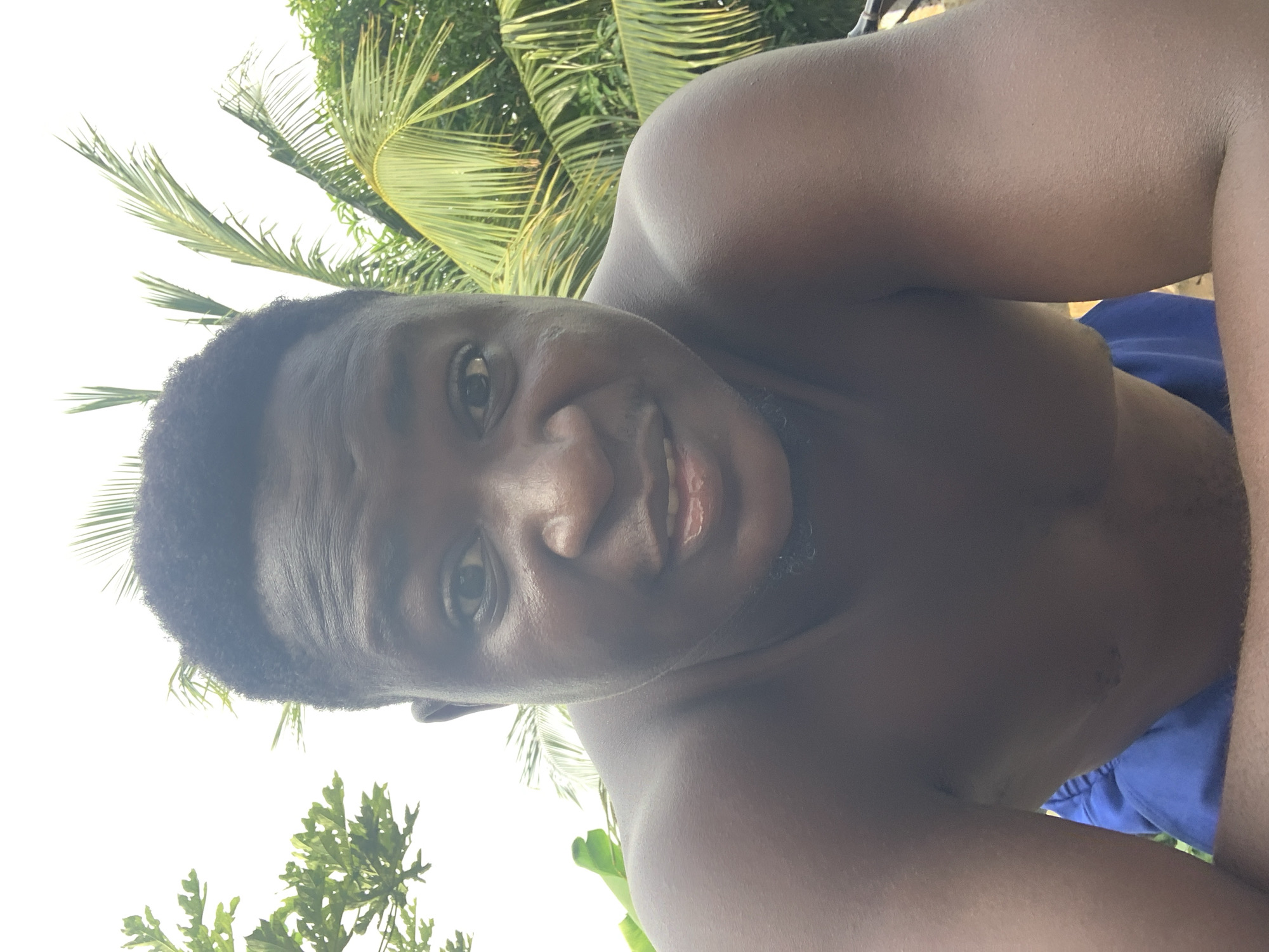 A selfie-style shot shows a Black male partially undressed, possibly wearing blue shorts, taken outdoors in Akwanga, Nigeria. Green foliage, including palm trees and other greenery, forms the background. The sky is partially visible in the upper left.