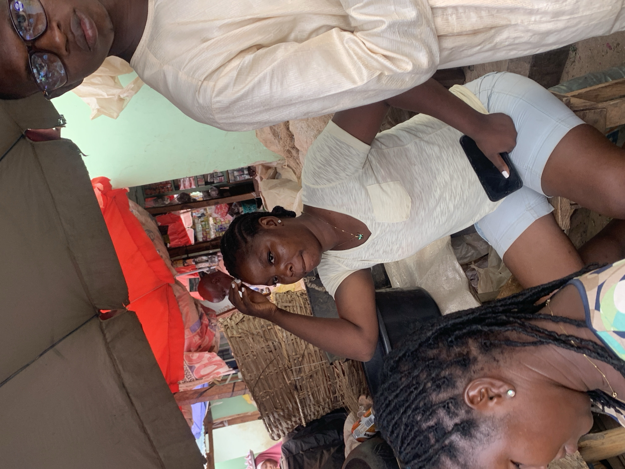 The image captures an active market scene in Akwanga, Nigeria, under partial canopy shade provided by grey and red tarps. In the central foreground, a dark-skinned woman with braided hair, wearing a light-colored, short-sleeved top with a chest pocket and light-colored shorts, is seated. She wears a gold necklace with a green pendant and holds a black smartphone in her right hand, her thumb extended. Her right arm is bent, with her hand positioned near her head. To her immediate right, a man with glasses and a light-colored, textured long-sleeved garment is partially visible, facing forward. In the immediate foreground left, the back of a person's head with black dreadlocks and a white stud earring is visible. The background features various market elements, including stacked woven baskets, a black plastic basin, and bags containing unknown dry goods. Further back, individuals are visible, including one person in a red head covering and another older man with a bald head. Market stalls with assorted products are discernible against a light green wall and other wooden structures.