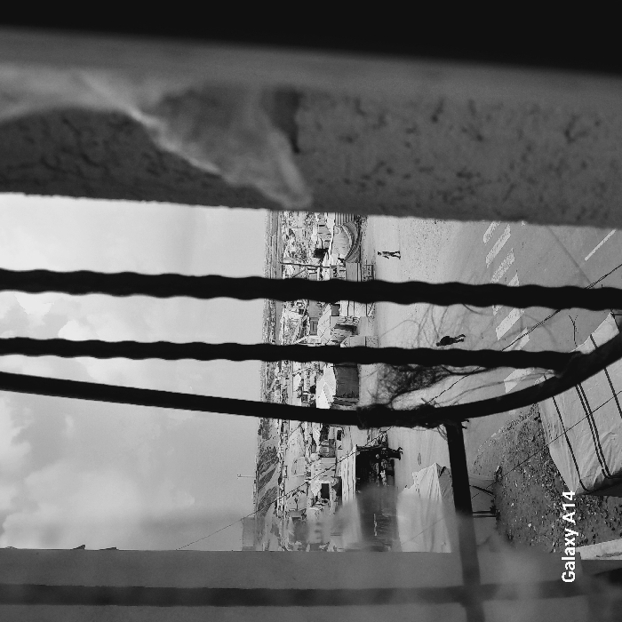 Captured from an elevated perspective through a wire fence, the black and white image features a town's landscape. Buildings, potentially residential, are visible, along with a street displaying pedestrian crossings. Two people are seen traversing the road. The composition includes a portion of a window frame at the top and a blurry foreground, adding depth to the urban scene. The text "Galaxy A14" is visible along the edge of the image.