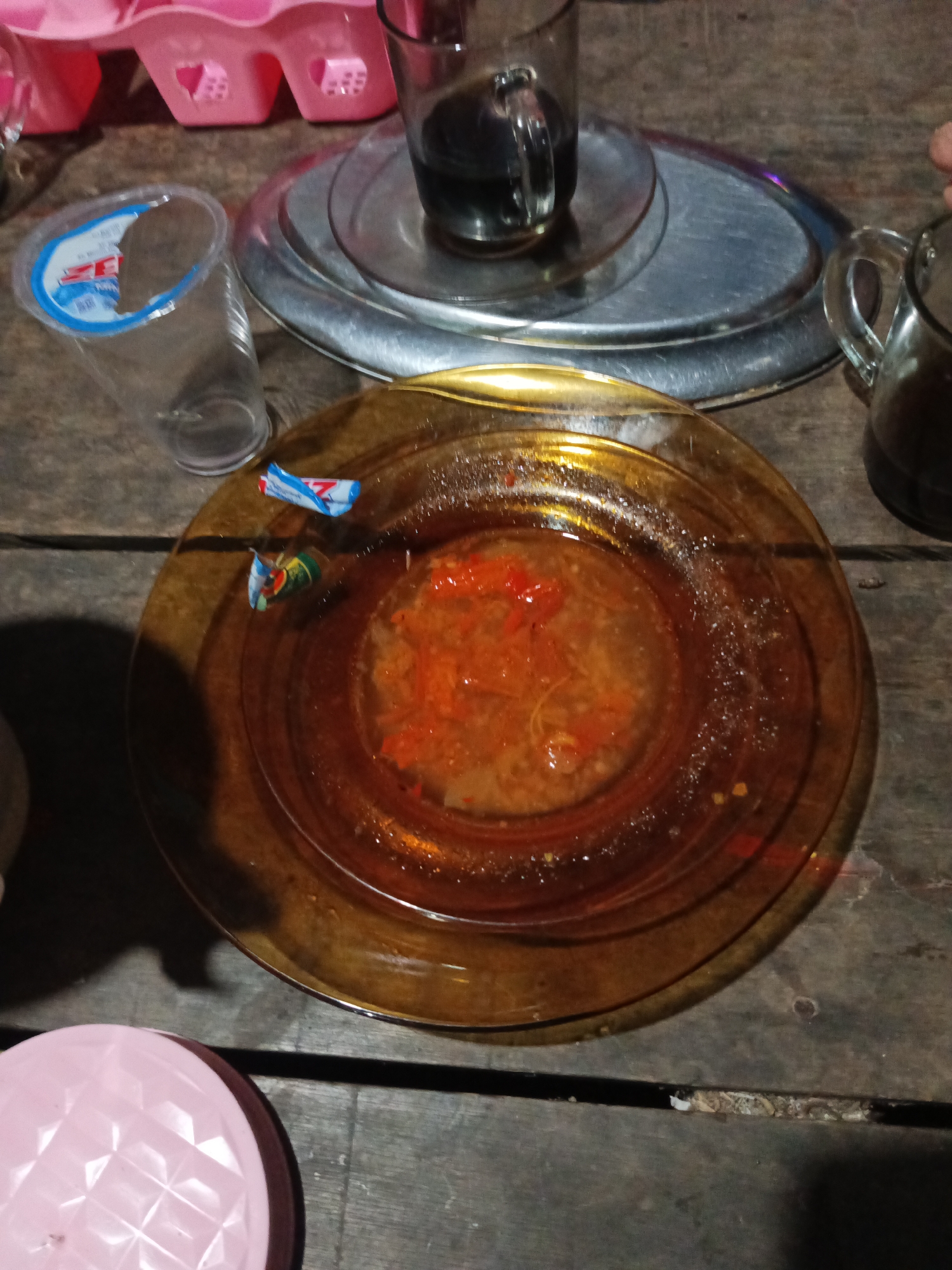 An overhead shot shows a rustic wooden table with various items on it, suggesting a casual meal setting. A clear plastic cup with a blue and white label is positioned to the left, partially filled with water. Next to it, a round, amber-colored plate holds a portion of what appears to be a saucy dish with visible red chili pieces and some fibrous material. Two small, colorful wrappers, one red, white, and blue, and another with green and yellow, lie on the plate. Behind the plate, a clear glass cup filled with dark liquid, likely coffee, sits on a round, metallic tray. Another glass cup with a handle, also containing a dark liquid, is visible to the right, partially obscured. In the background, a pink plastic tray with heart-shaped compartments is present. The scene is illuminated by overhead lighting, casting shadows on the table. The overall impression is that of a simple, everyday meal or snack, possibly in a rural or informal environment.