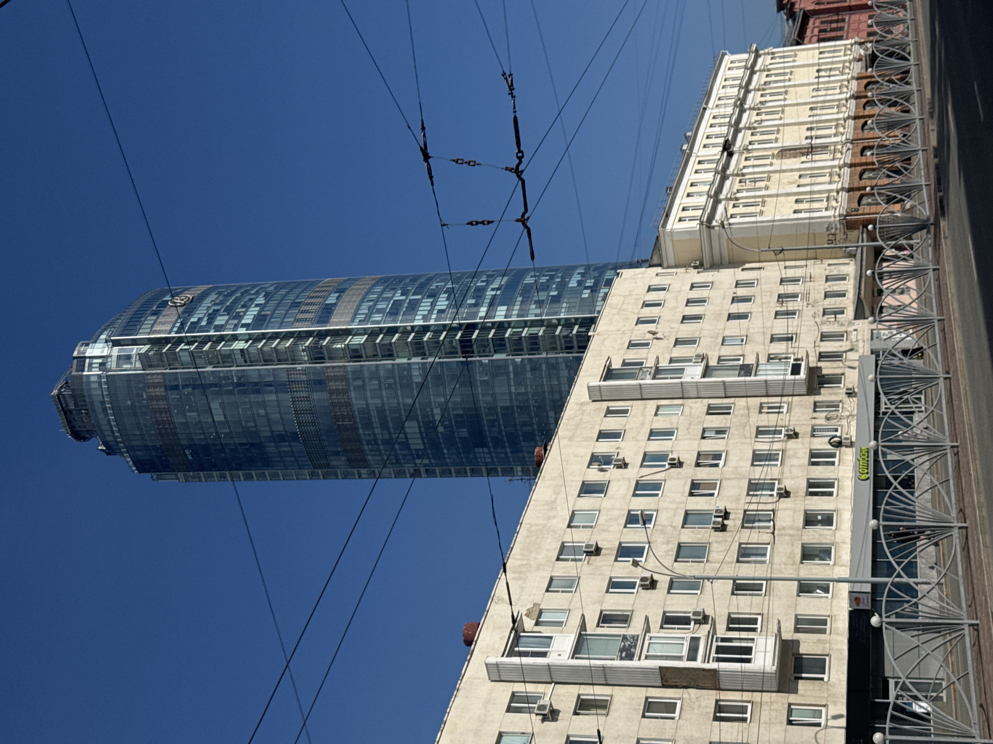 The image depicts a street scene in Yekaterinburg, Russia, dominated by a tall, modern glass skyscraper. In the foreground, a long, multi-story building with a light-colored facade is visible, featuring numerous windows and balconies. Several air conditioning units are mounted on the facade. Below this building, a street-level commercial area with a gray awning and the word "СОТПРО" (SOTPRO) in yellow lettering is partially obscured by a decorative metal fence. The fence, with a recurring fan-like pattern, runs along the sidewalk. Overhead, complex network of electrical wires, likely for public transportation, crisscross the sky. The sky is a clear, deep blue. In the background, a slender, glass-clad skyscraper with a rounded top stands tall, reflecting the blue sky and surrounding buildings. The skyscraper has a distinctive pattern of darker bands on its facade. To the right of the main foreground building, a narrower, lighter-colored building with a more traditional architectural style, featuring arched windows on the lower level, is visible. Further to the right, the corner of a red brick building is visible. The overall impression is an urban environment with a mix of older and contemporary architecture.