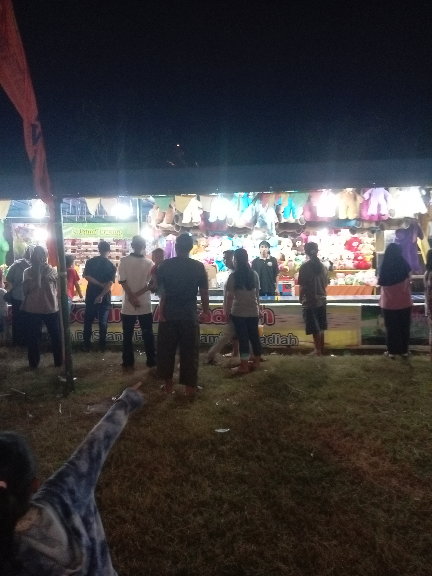The image depicts an outdoor night scene at what appears to be a carnival or amusement fair in Batam, Indonesia. In the background, a brightly lit stall is visible, adorned with colorful stuffed animals hanging as prizes. A banner above the stall reads "Selamat Datang" (Welcome). Several people are gathered around the stall, some looking at the prizes, while others stand and interact.
In the foreground, a person's arm and shoulder are visible, pointing towards the left side of the frame. The ground is covered in grass, and debris is scattered in some areas. The overall atmosphere suggests a lively public event taking place after dark.