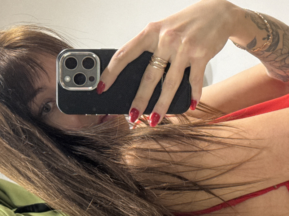 A person holds a black smartphone, obscuring part of their face, with the camera lenses visible. Their long brown hair frames the side of the phone. The person's hand, adorned with three gold rings and glossy red nails, grips the device. Part of a gold bracelet and tattoo are visible on the wrist. They are wearing a red garment. The overall perspective suggests a selfie taken in a casual setting.