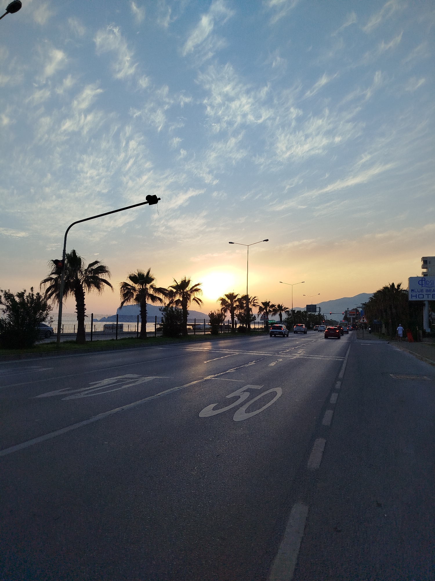 The image captures a street view in Alanya, Turkey, at sunset. The sky is filled with cirrus clouds and transitions from blue to orange near the horizon, where the sun is setting. Palm trees line the left side of the road, obscuring the view of the beach and ocean. A silhouette of a mountain is visible in the distance.
The road is marked with a speed limit of "50" painted on the surface, and cars are driving on the road. Street lights illuminate the scene. On the right side of the road, there is a sign for the "Blue Beach Hotel." A person is walking on the sidewalk. The scene appears to be taken during the evening with the sun setting.