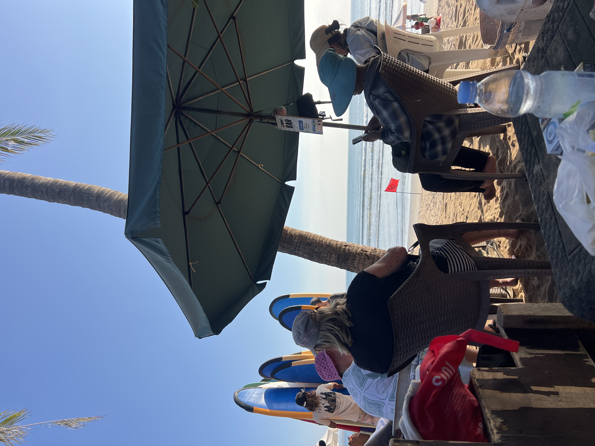The image depicts a beach scene in Kuta, Indonesia, with several people seated under a large green umbrella, facing the ocean. In the foreground, a wooden structure holds a red bag. A plastic water bottle and a pack of cigarettes are on a table in front of the seated individuals. Two people are looking at their phones. Behind them, several surfboards stand upright on the sand. A palm tree trunk is visible to the right of the umbrella, and the ocean with gentle waves stretches to the horizon under a clear blue sky. A red flag is planted in the sand to the left of the umbrella. The overall atmosphere is relaxed, with people enjoying a day at the beach.