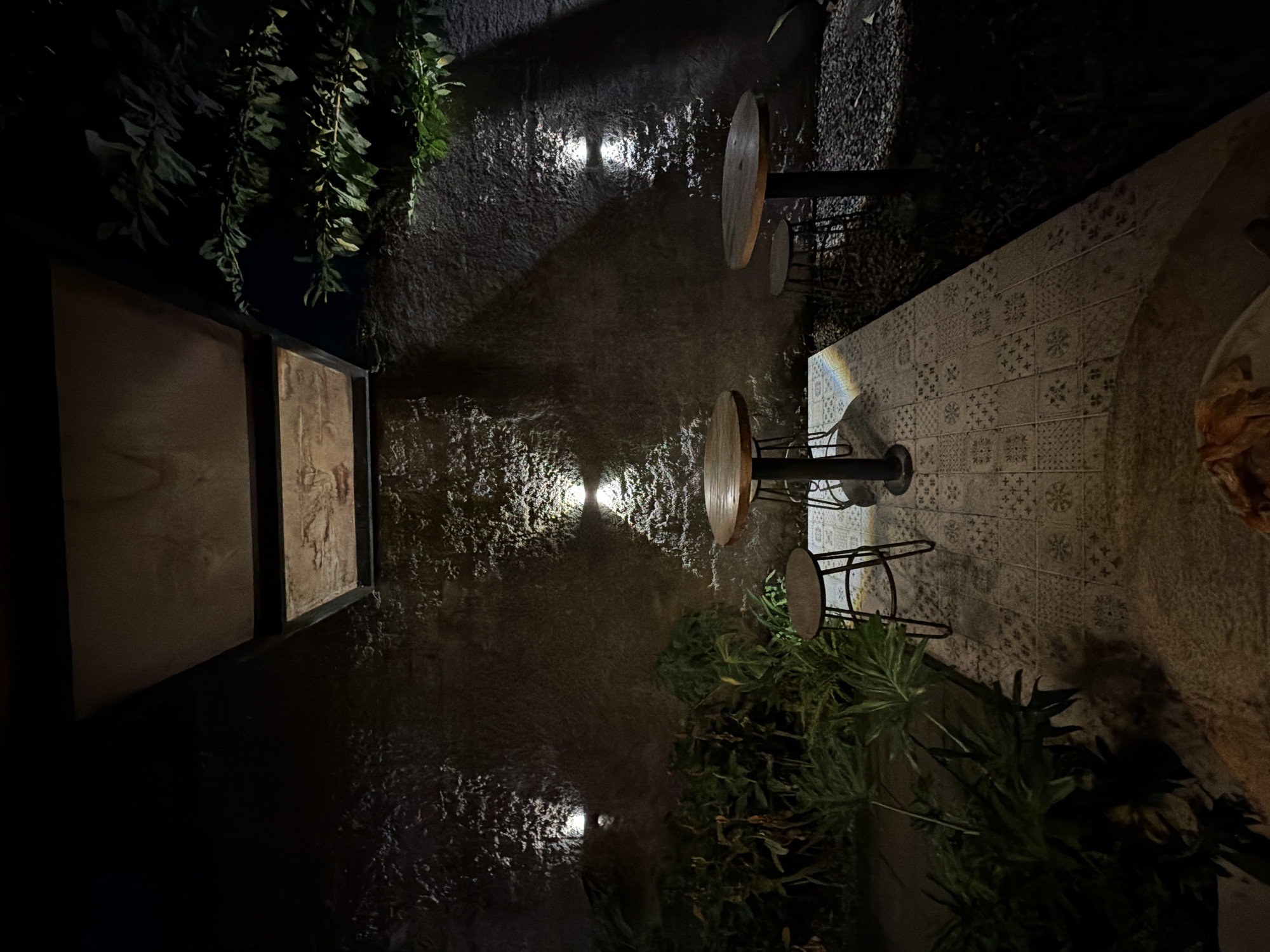 The image depicts an outdoor patio area at night, possibly a cafe or restaurant, in North Kuta, Indonesia. The scene is dimly lit by several wall-mounted spotlights that illuminate a textured, dark grey wall. In the foreground, a tiled patio floor is visible, interspersed with gravel. Two round, wooden-topped tables with dark metal bases are present, accompanied by two dark metal stools. Lush green foliage can be seen to the left of the tables, suggesting a garden or planted area. A partially visible overhead structure, possibly a roof or awning, is also present. There are no people visible in the image.