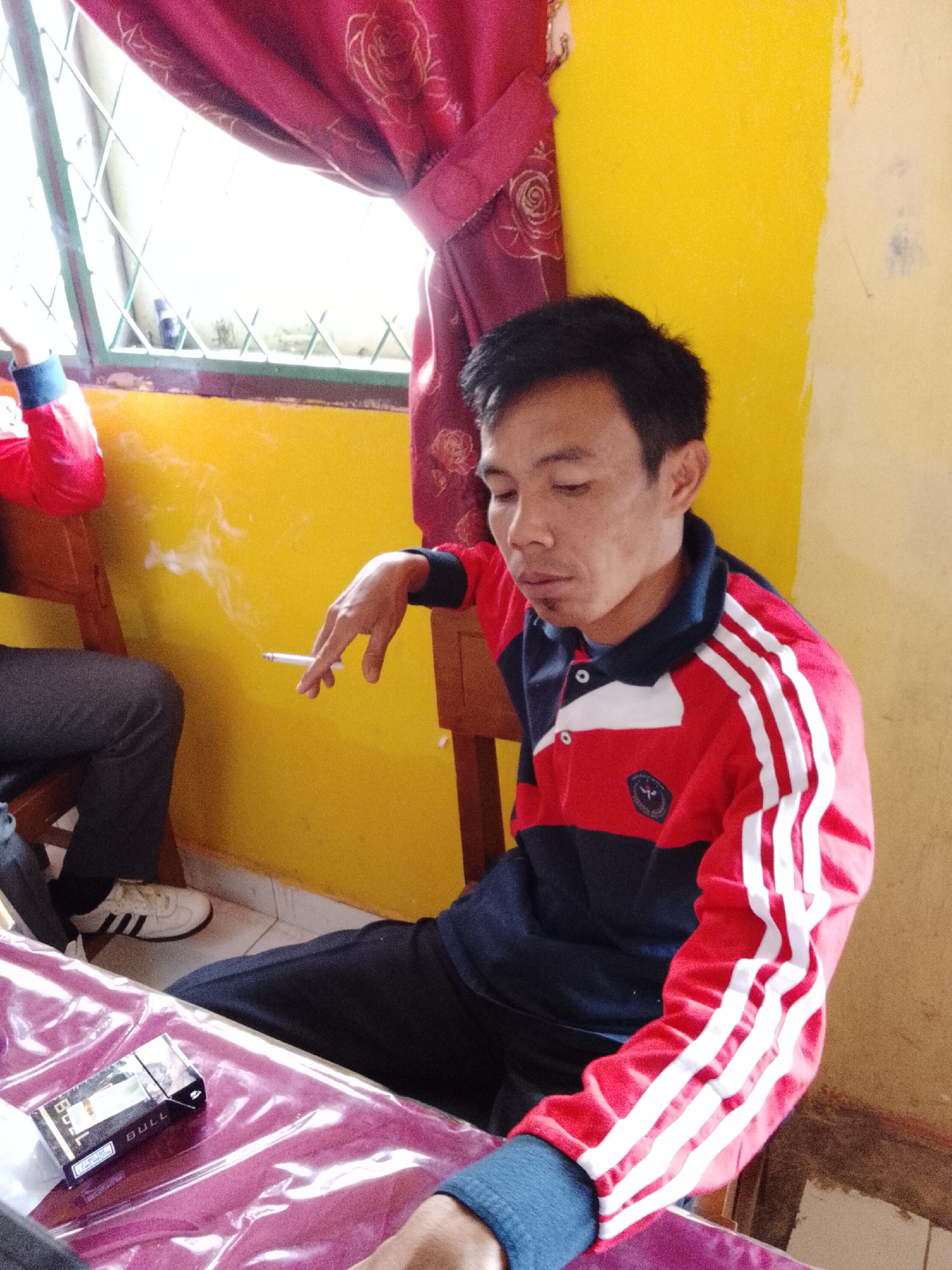 A man in a red and blue collared shirt with white stripes on the sleeves is sitting at a table in what appears to be a home or small shop in Jepara, Indonesia. He is holding a lit cigarette and exhaling smoke. A pack of "BULL" brand cigarettes is on the table in front of him. The room is painted yellow and has red curtains. There is a window with a lattice pattern in the background. The man is wearing black pants and white sneakers.
