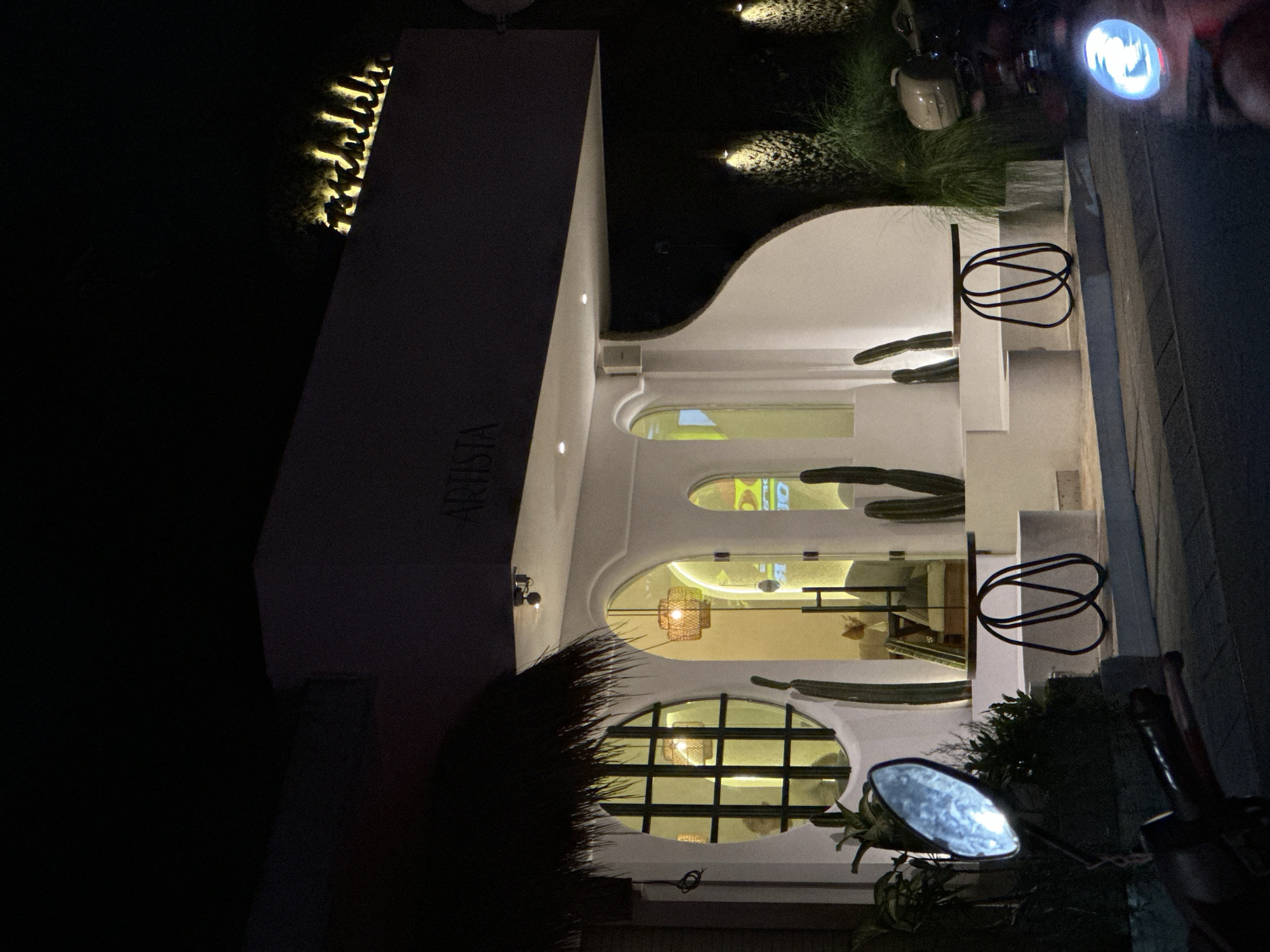The image shows a building named ARISTA in Canggu, Indonesia, at night. The building is predominantly white with arched windows and unique decorative elements, including what appear to be cacti integrated into the design. Light emanates from within the building, illuminating its interior and exterior facade. The building features a modern architectural style with clean lines and minimalist elements. To the side of the building, a motorcycle is partially visible. Vegetation surrounds the building, adding a touch of natural contrast to the modern architecture. The surroundings appear dark, typical of a night scene.
