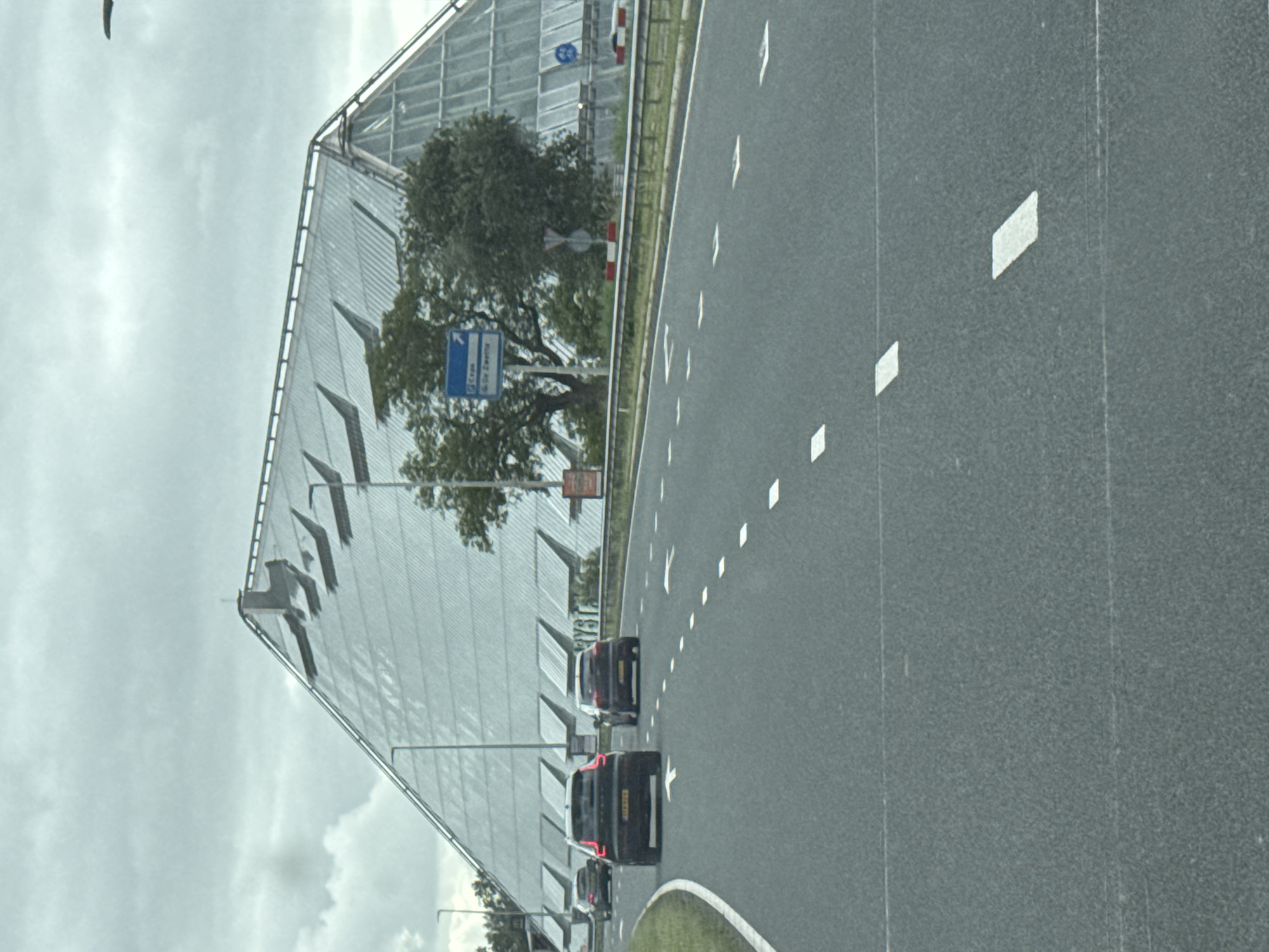 The image depicts a grey, overcast day with a modern, pyramid-shaped building prominently featured. The building has a distinctive faceted roof made of glass and metal, with numerous angled panels that appear to be adjustable skylights or ventilation openings. The structure is large and architectural, possibly a public building or a corporate headquarters. In the foreground, a multi-lane asphalt road curves to the left. Several cars are visible on the road, including a black SUV in the right lane of the foreground and a dark-colored sedan in the left lane. Road markings, including white dashed lines and directional arrows, are clearly visible. A blue road sign with white text and an airplane icon is situated to the left of the road, indicating directions to "Copa" and "De Zeelte." Trees and greenery are visible behind the road and surrounding the building. The overall scene suggests a location along a major roadway near a significant architectural landmark in Leeuwarden, Netherlands.