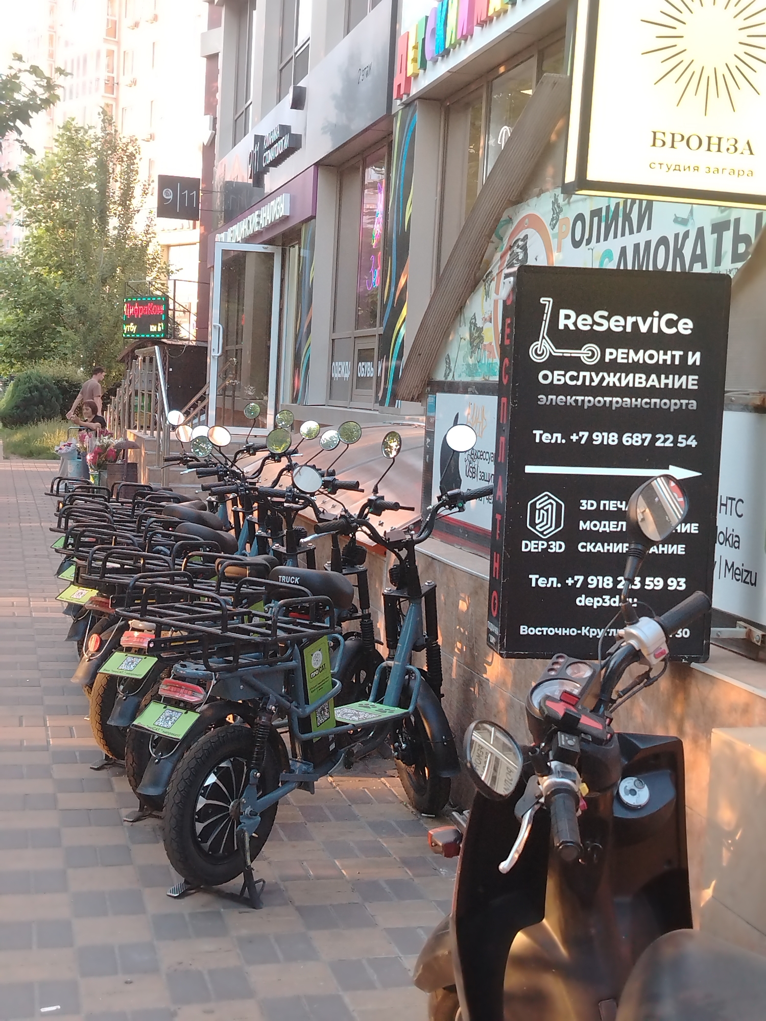 The image shows a row of electric scooters parked on a sidewalk in front of a building with several storefronts. The building has a modern architectural style with large windows and signage. One prominent sign displays "ReService" with information about scooter repair and maintenance, including phone numbers and services like 3D printing and scanning. Below this, there's a smaller sign indicating "Vostochno-Krugaya."
In the background, several people are visible. Two individuals, a man and a woman, are standing near a small stall with flowers, possibly making a purchase. Another person is walking away from the camera on the sidewalk. The environment suggests an urban setting with paved sidewalks and trees lining the street. The lighting indicates it's either early morning or late afternoon, with soft shadows and warm sunlight.
One of the electric scooters in the foreground has the word "TRUCK" printed on its rear rack. The scooters are equipped with storage baskets and have distinct green tags on the rear fenders, likely indicating rental services. The overall scene depicts a common urban amenity – electric scooter rental and service – in a commercial district.