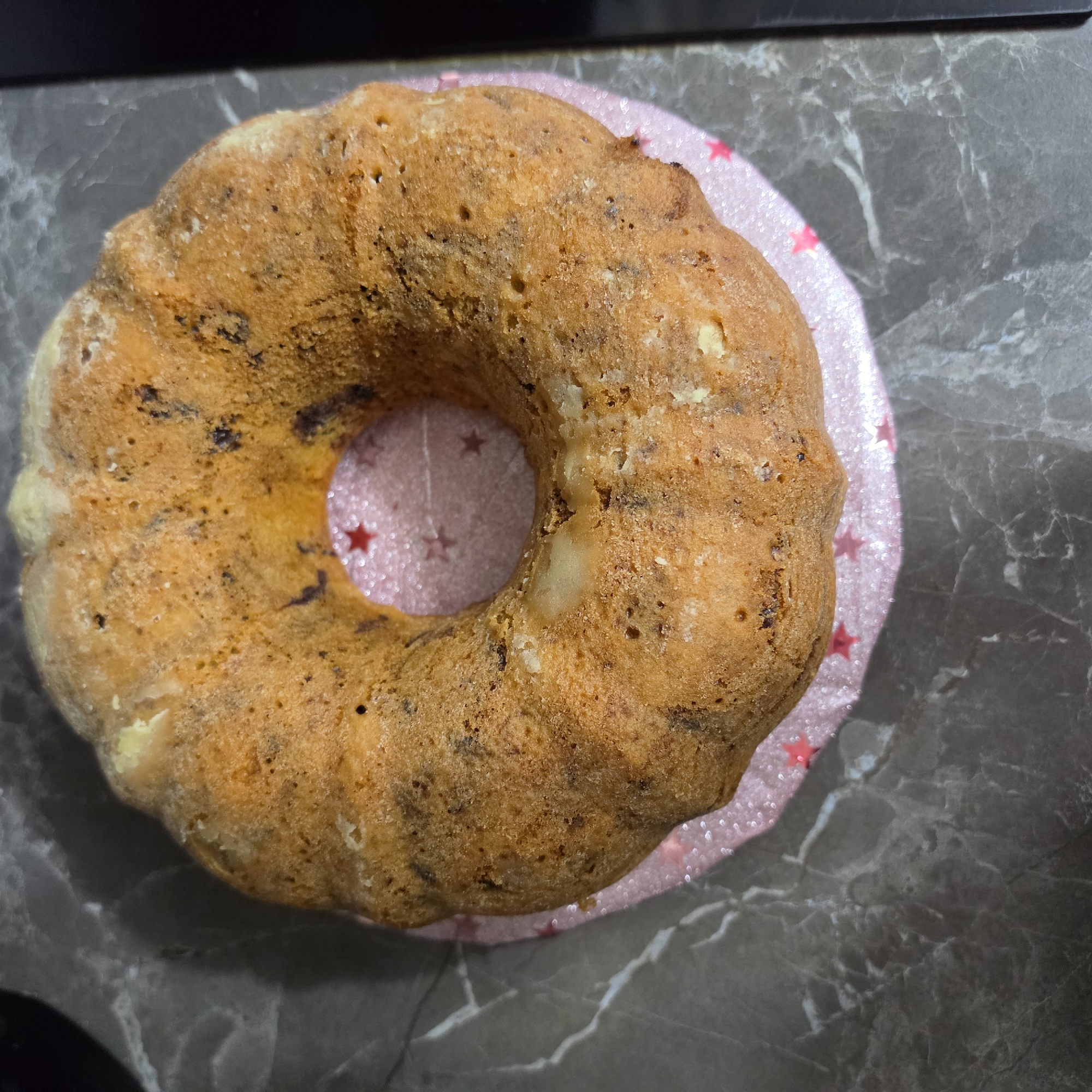 A bundt cake with chocolate chips rests on a pink star-patterned plate, which is on a dark gray marble countertop. The cake is golden brown with visible chocolate chips and some lighter, possibly undercooked, spots. The background is a dark gray, marble-like surface with some lighter grey veining. There are no people or signs of the city of Peshtera, Bulgaria in the image.