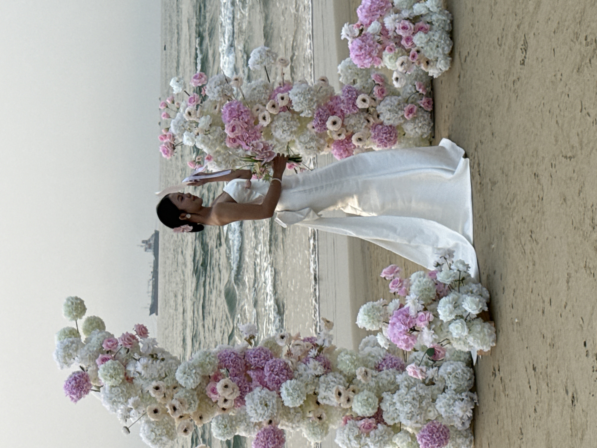 A woman in a white strapless dress stands on a beach, holding a bouquet of flowers. The background shows the ocean and a blurred ship in the distance. An archway made of pink and white flowers surrounds her. The image was likely taken in Đà Nẵng, Vietnam, given the beach setting.