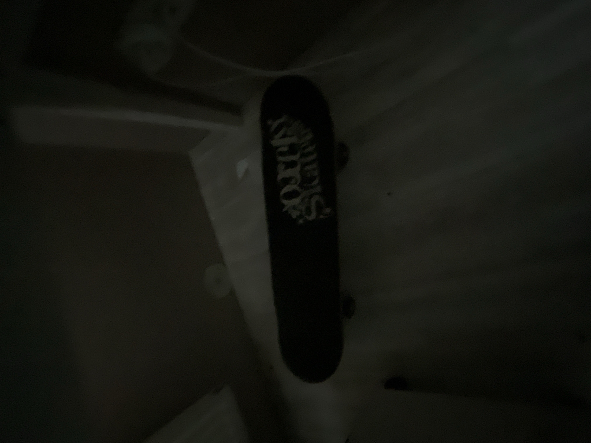 A black skateboard with a white logo is resting on a dark, possibly carpeted, surface. The logo on the skateboard appears to read "Only Skates" in a stylized font. The background is dark and indistinct, suggesting a dimly lit indoor environment, possibly a room. There is no discernible urban or natural landscape visible that would indicate a specific location within Мухавецкі сельскі Савет, Belarus. The image is of low quality due to darkness and potential motion blur.