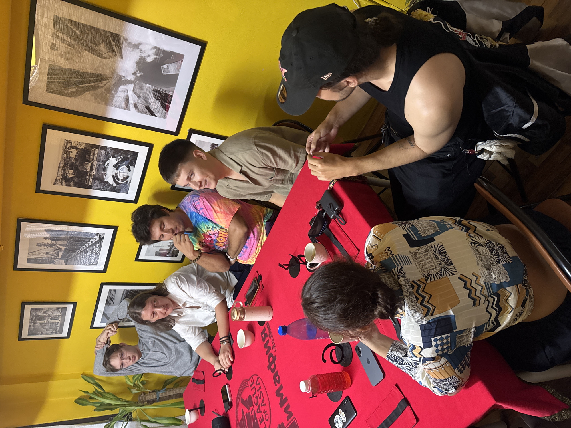 A group of six individuals are gathered around a long red table, seemingly for a podcast or discussion. The room has bright yellow walls adorned with several framed black and white photographs. On the table are microphones, cups, bottles of water and a red beverage, and mobile phones. The individuals are dressed casually. One person in the foreground on the right wears a black baseball cap and a black tank top, and appears to be speaking into a microphone. Another person on the left wears glasses and a grey hooded sweatshirt. In the center of the table, a woman in a white shirt is seated, and beside her, a man in a tie-dye shirt gestures with his hand. To the right of him, a man in a light-colored collared shirt is looking down. In the foreground, another person with dark hair is looking down at the table, wearing a patterned shirt. The room also contains a tall potted plant on the left side of the image. The setting is an indoor space.
