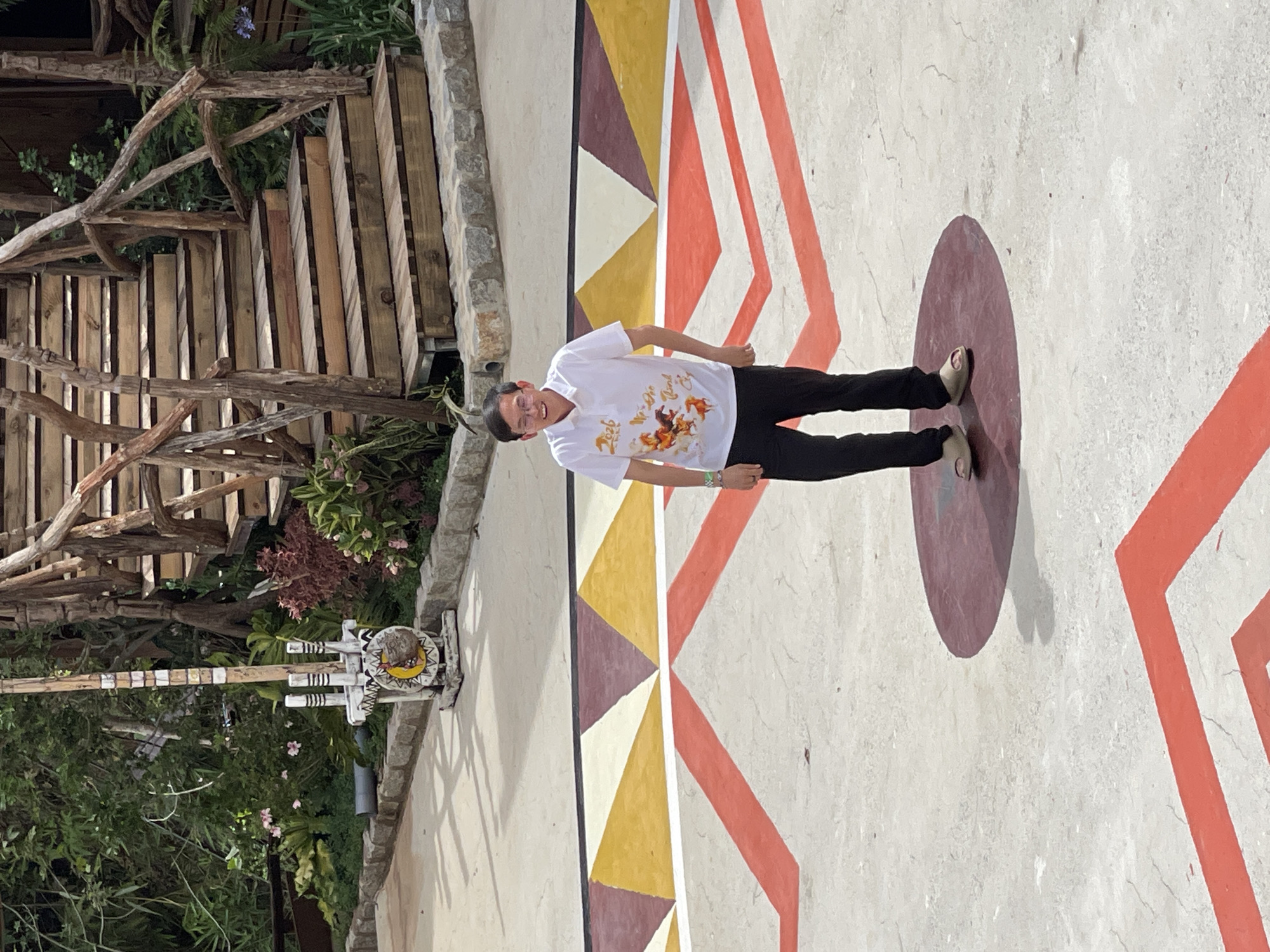 The image depicts a person standing on a painted circle on a patterned ground in Thôn 5, Vietnam. The person wears a white shirt with a graphic, dark pants, and sandals. The patterned ground features triangles and broad red lines. To the left, there's a wooden structure and greenery. A tall, possibly decorative, post stands near the edge of the frame, closer to the wooden structure.