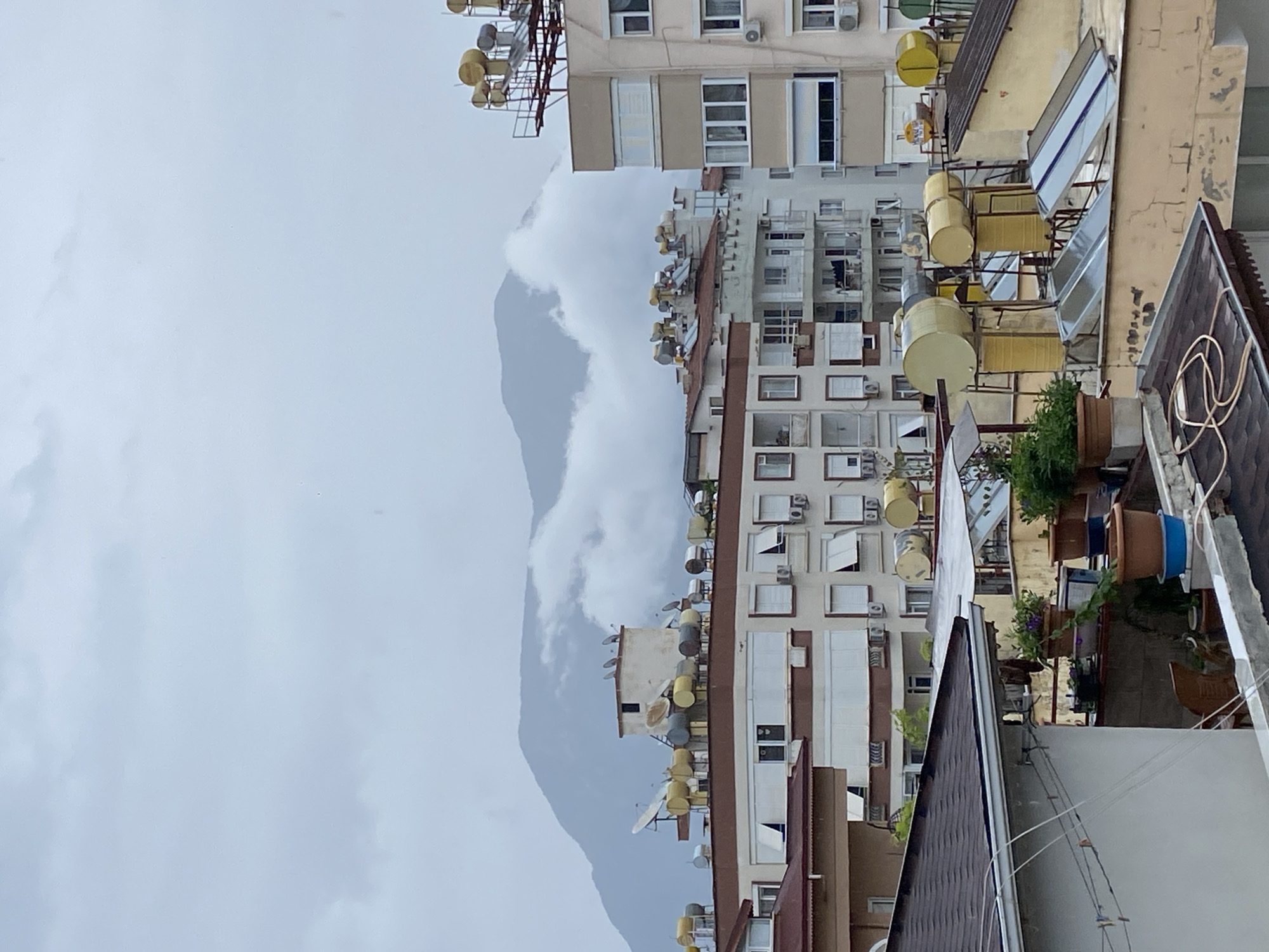 The image depicts a cityscape in Alanya, Turkey, under an overcast sky. In the foreground, there are rooftops of buildings. The middle ground shows densely packed apartment buildings with numerous windows, some with air conditioning units. Many of the buildings are topped with solar water heaters or satellite dishes. The background features a mountain partially obscured by clouds. The overall impression is one of a dense urban environment in a coastal setting.