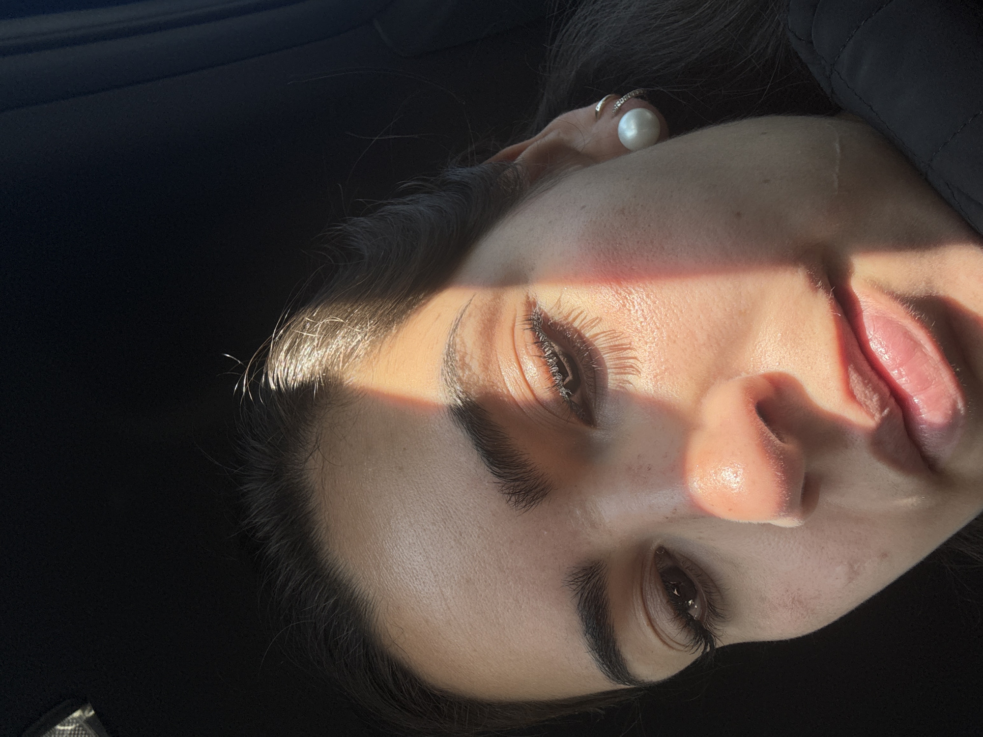 A close-up portrait of a woman's face, tilted to the right. She has dark hair and is wearing a pearl earring and a ring. Light shines across her forehead and nose. The background is dark. This is in the city of Puebla, Mexico.
