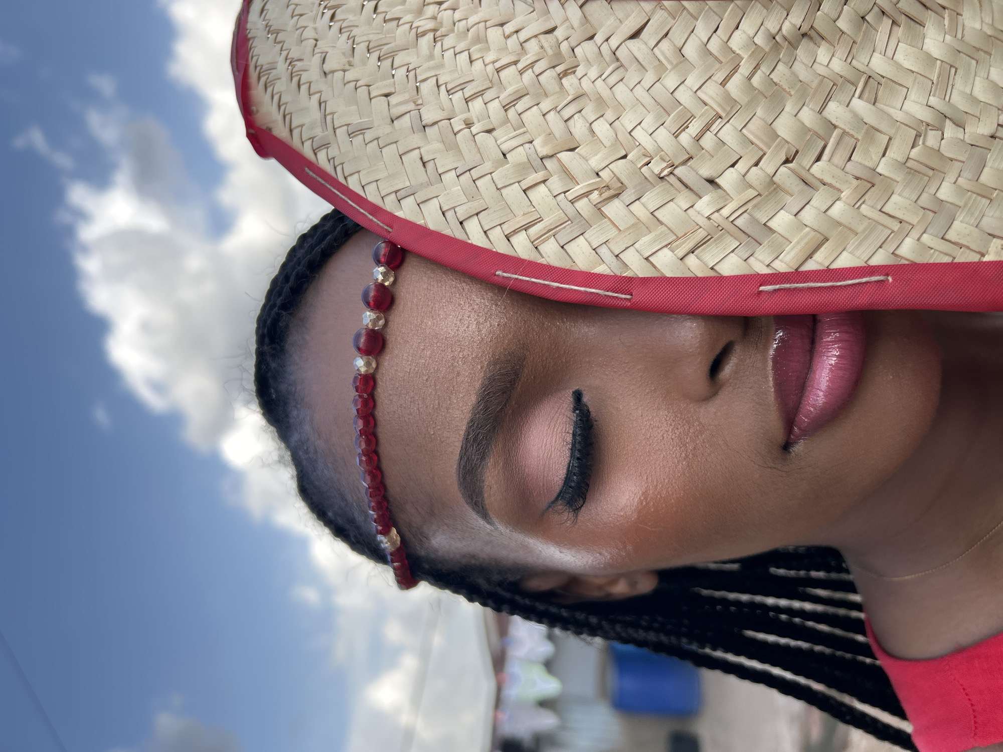 This is a close-up, vertically oriented photograph of a young Black woman wearing a straw hat with a red band. She has braids and is wearing a beaded headband with red and gold beads. Her eyes are closed, and she has dramatic eye makeup with dark eyeliner and false eyelashes, along with shimmery pink eyeshadow. Her lips are glossy and painted a shimmery pink.
The background is a bright blue sky with some white clouds, suggesting it is daytime. In the blurred background, there are hints of structures that might be in a residential area, including what looks like laundry hanging on a line and some blue barrels. The setting appears to be outdoors, likely in a warm climate, given the presence of the straw hat.
The overall mood of the image is one of relaxed confidence or peaceful contemplation. The lighting suggests it is either morning or afternoon, with bright sunlight illuminating the subject's face. The image was taken in Jos, Nigeria. There is no visible text in the image.