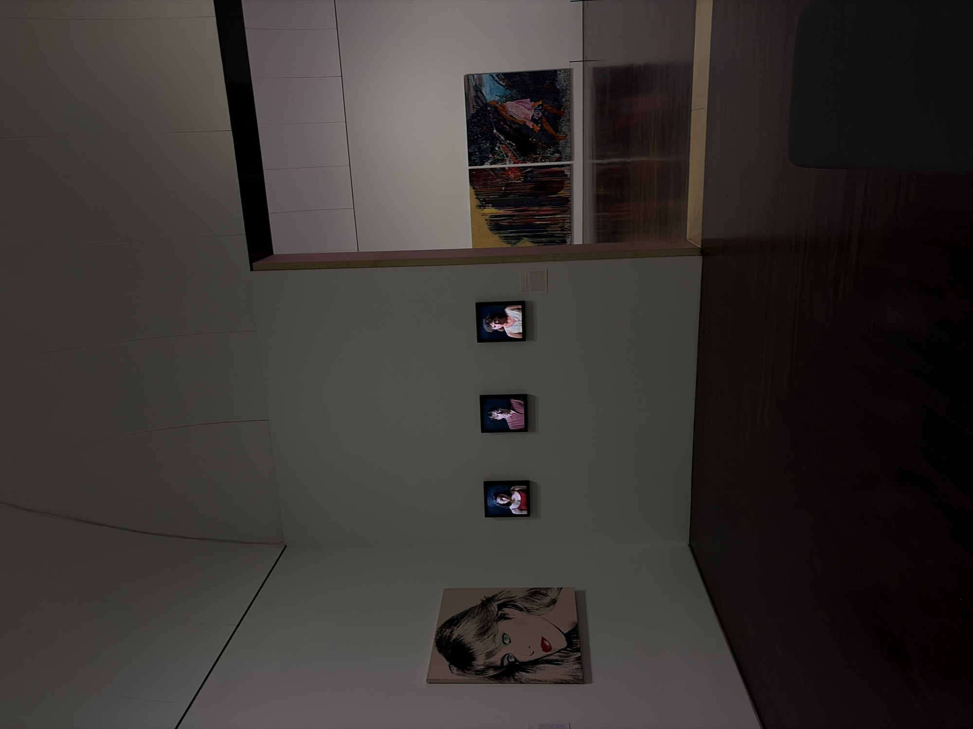 The image shows an interior space with artwork displayed on the walls. On one wall, there are three small framed portrait photographs hanging vertically above a larger canvas depicting a woman with blonde hair and red lips in a pop art style. On an adjacent wall, there are two abstract paintings stacked one above the other, appearing as two halves of a larger work. The room seems to be a gallery or exhibit space, with neutral-toned walls and a light wood-look floor. The lighting in the image is subdued, creating shadows and emphasizing the artwork. The scene suggests an art exhibit. There is no indication of the location in the city of Austin, United States of America.