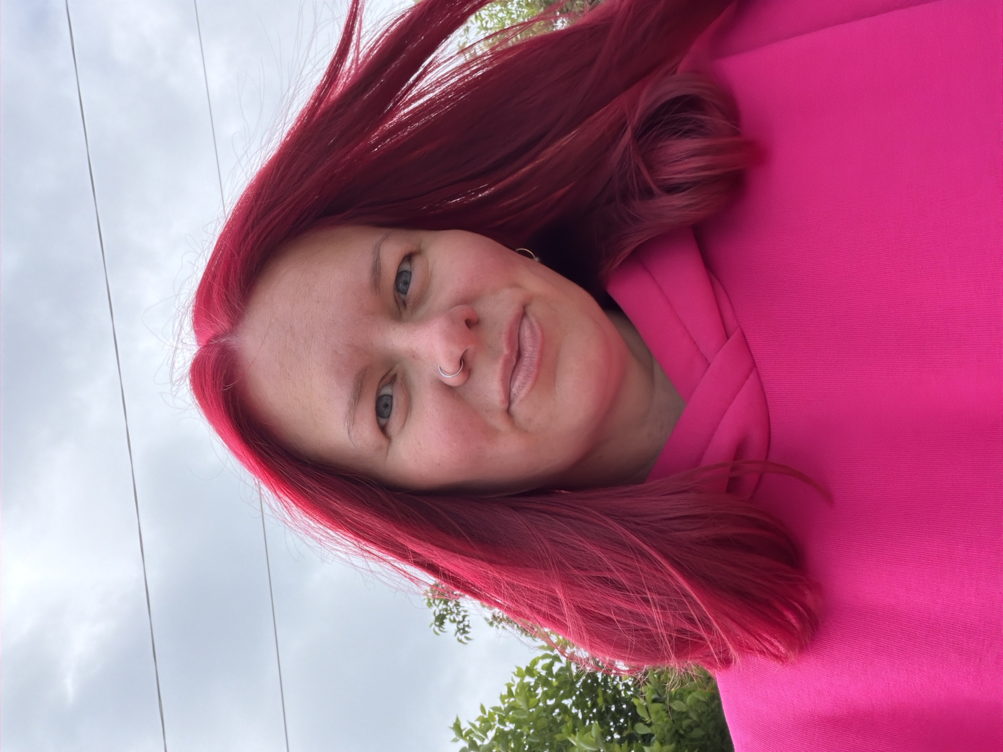 The image is a vertical, close-up outdoor portrait of a person with vibrant, bright red hair. The individual is wearing a fuchsia or bright pink hooded sweatshirt and is looking directly at the camera with a gentle smile and a calm expression. They have light skin, visible blue eyes, and a silver hoop nose ring in their left nostril.
The photograph is taken from a low angle, with the person's head significantly tilted, giving the composition a dynamic feel. The background primarily consists of a bright, overcast sky, indicating daytime and cloudy weather. Overhead power lines are visible against the sky in the upper left portion of the image. Green foliage, likely from trees or bushes, can be seen in the lower right, suggesting an outdoor setting such as a park or street in Chaikovsky, Russia. No text is visible in the media file.