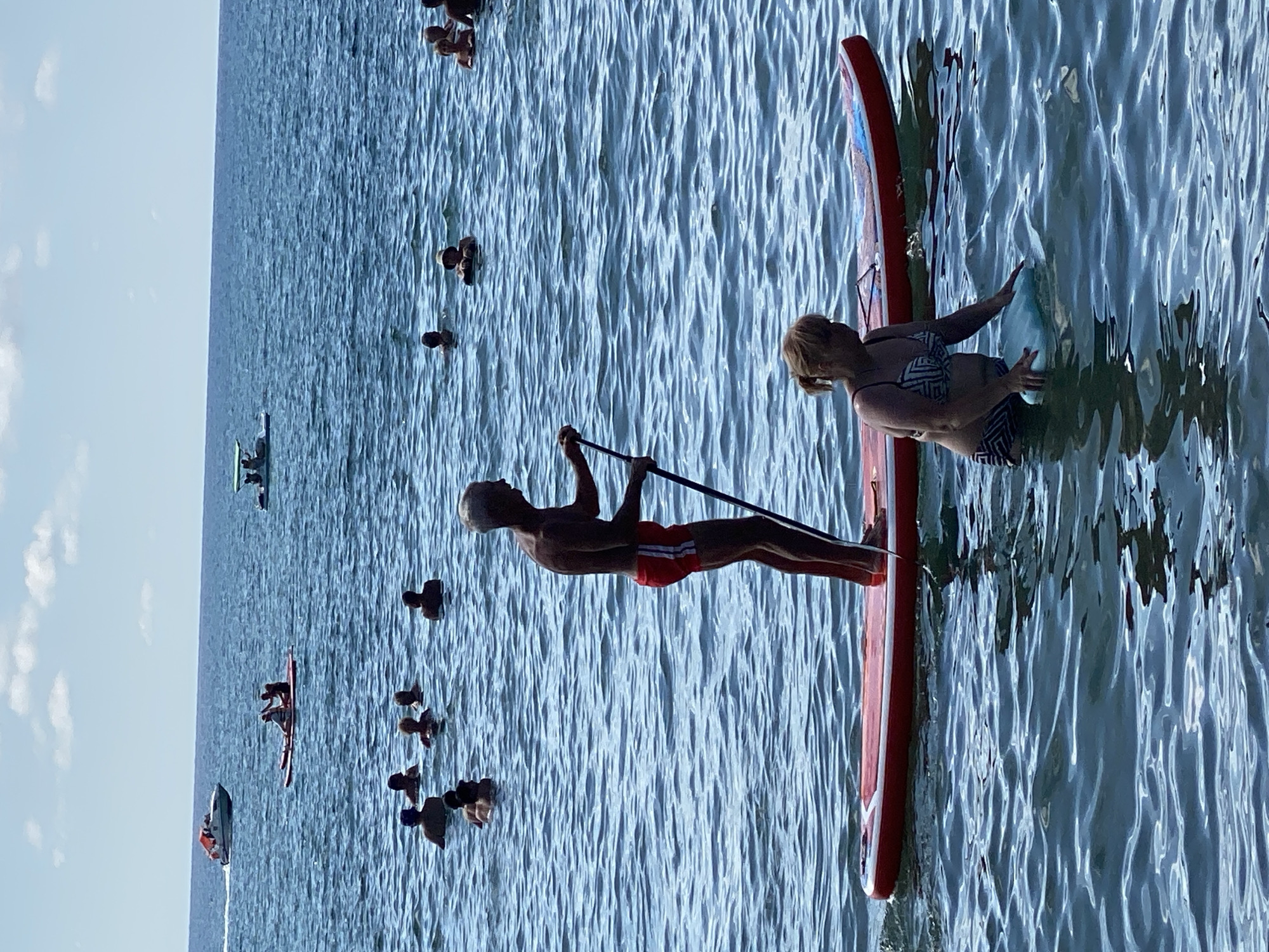 In Arkhipo-Osipovka, Russia, a man with gray hair stands on a red paddleboard, holding a paddle. He is wearing red swim trunks. A woman in a patterned bikini stands in the water next to the paddleboard, holding a light blue object. Several other people are visible in the background, some swimming and some on other watercraft. The scene is set on a calm, blue sea under a partly cloudy sky.