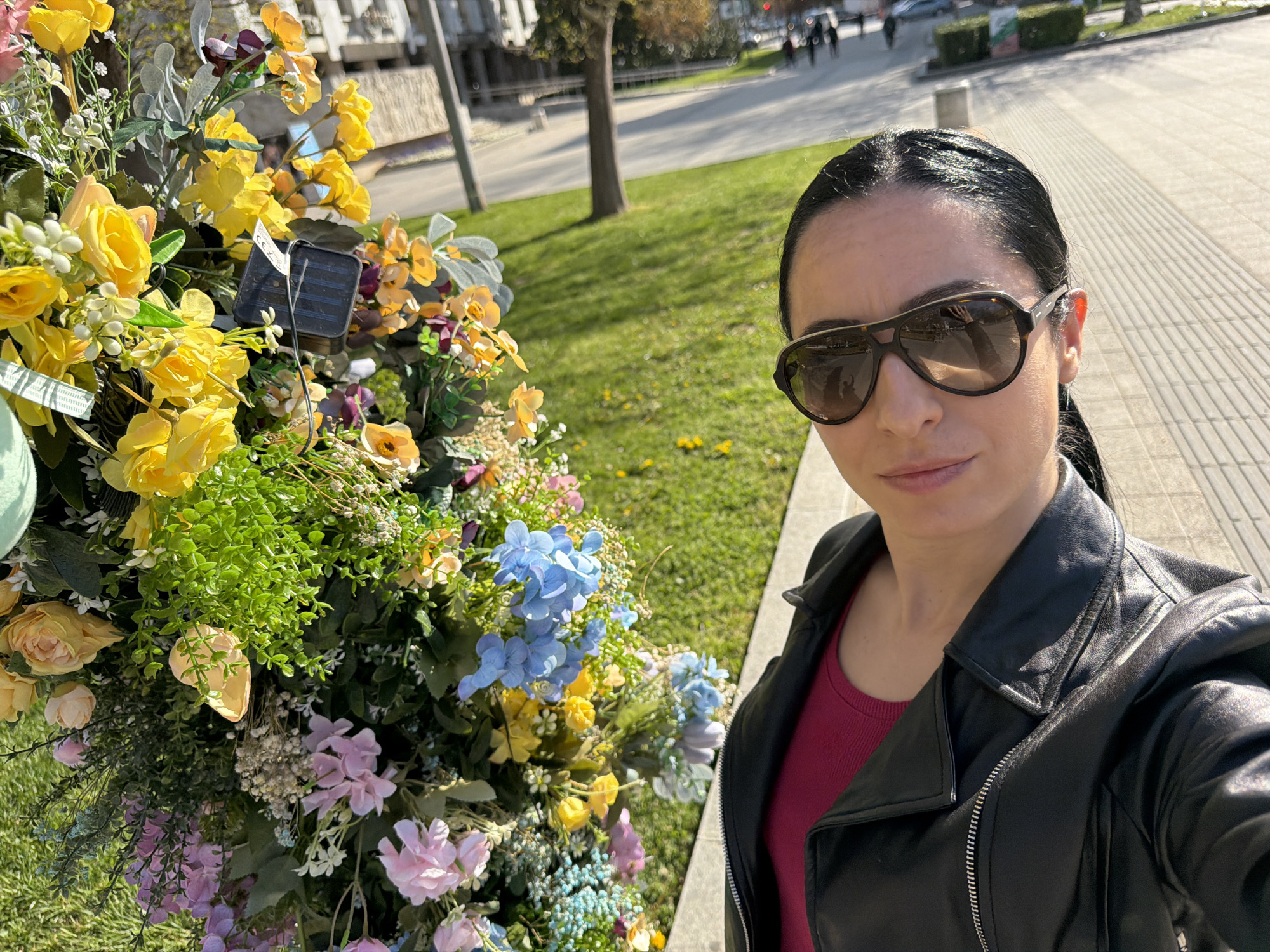 A woman takes a selfie outdoors in a sunny, daytime setting, likely in Pleven, Bulgaria. She is positioned to the right of the frame, wearing large, dark sunglasses, a black leather jacket, and a dark red top. Her dark hair is pulled back, and her expression is neutral.
To her left and filling the foreground is a vibrant display of artificial flowers and greenery. Prominent yellow roses, light blue hydrangeas, and various shades of pink and purple blossoms are interspersed with rich green foliage. A small, black solar panel is visible nestled within the floral arrangement, connected by a thin wire. A tiny white tag with illegible text can also be seen.
The background shows an urban or park-like environment under clear skies. To the left of the woman, beyond the flowers, is a grassy area with scattered trees, leading to distant buildings and what appears to be a street with people walking. To her right, a paved sidewalk with a distinctive striped pattern stretches into the distance, with more buildings and distant figures visible. The bright sunlight casts clear shadows, indicating good weather.