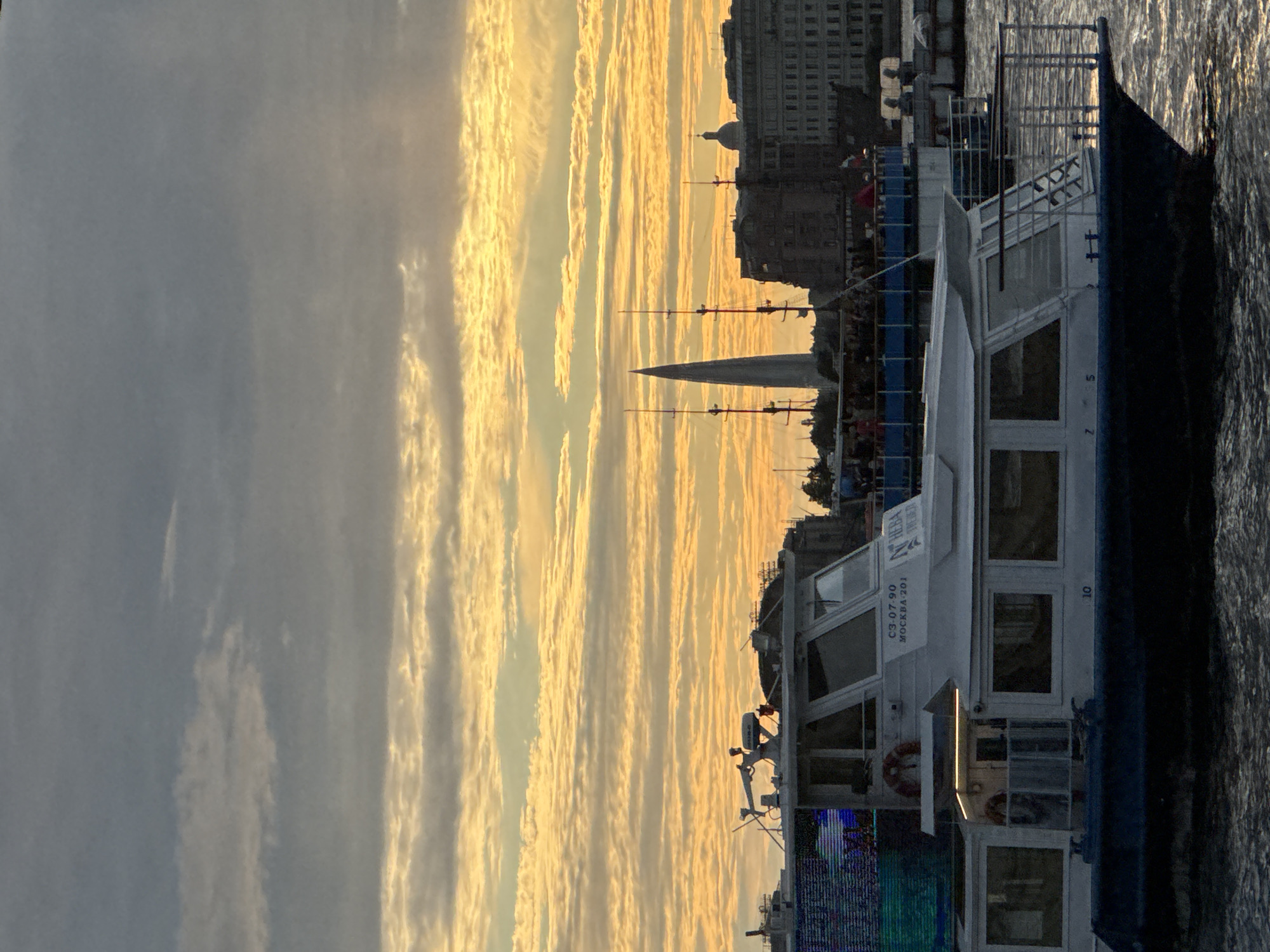 This media file captures a sunset over the Neva River in Saint Petersburg, Russia. The sky is filled with dramatic, elongated clouds tinged with vibrant golden and orange hues, suggesting the sun is low on the horizon. The water's surface reflects the warm colors of the sky, creating a shimmering effect.
Dominating the right side of the frame is a white passenger boat, likely a tour boat, docked or moving slowly on the river. The boat has multiple decks, large windows, and a dark hull. Visible on the side of the boat is the text "Neva" and "МОСКВА-201", indicating its name and possibly a designation or route. There is also a number "10" visible on the hull. A life preserver ring is mounted on the side of the boat.
In the background, the cityscape of Saint Petersburg is silhouetted against the colorful sky. Buildings with distinct architectural features, including a dome, are visible. A tall, pointed spire or monument also stands out against the horizon. The overall scene is serene, with the warm glow of the sunset creating a peaceful and picturesque atmosphere. The presence of the boat suggests a tourist or leisure activity is underway, taking advantage of the scenic evening light. The weather appears clear, with only the clouds contributing to the visual drama.