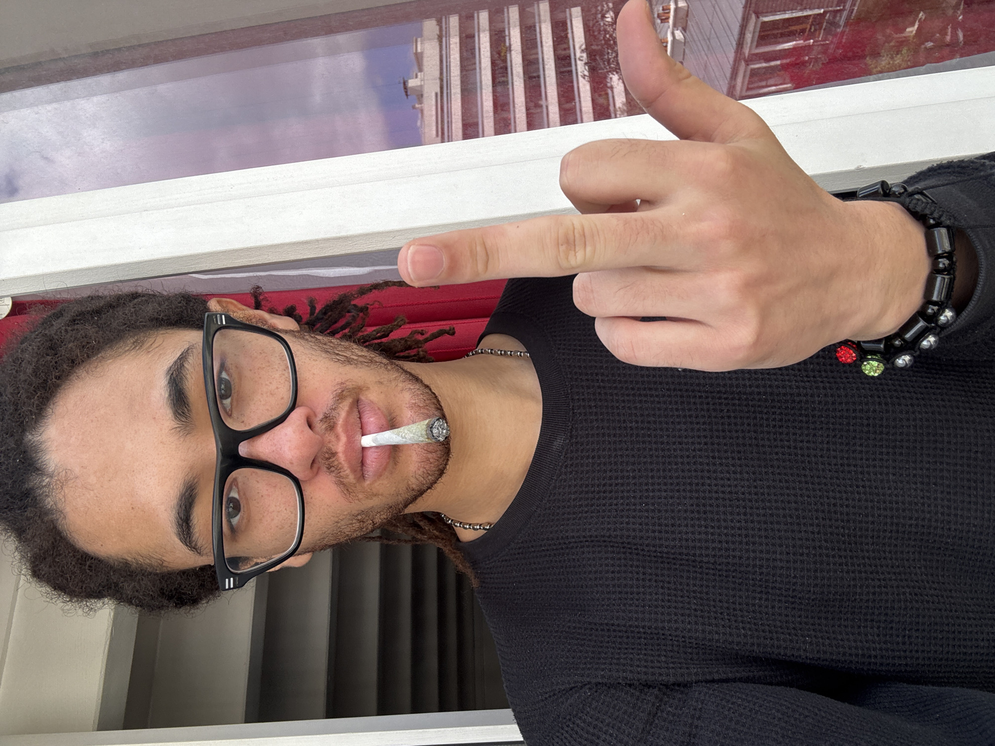 A young adult male, light-skinned, with dreadlocks and wearing eyeglasses, is posing indoors, holding a lit cigarette in his mouth. He is wearing a black waffle-knit shirt and a beaded bracelet. The background shows a window frame, with the reflection of buildings visible through the glass, potentially suggesting an urban environment. The reflection appears to show light-colored buildings with a red accent on one. The subject is holding up a finger to the camera. There is no definitive evidence that this is the city of Paris, France.