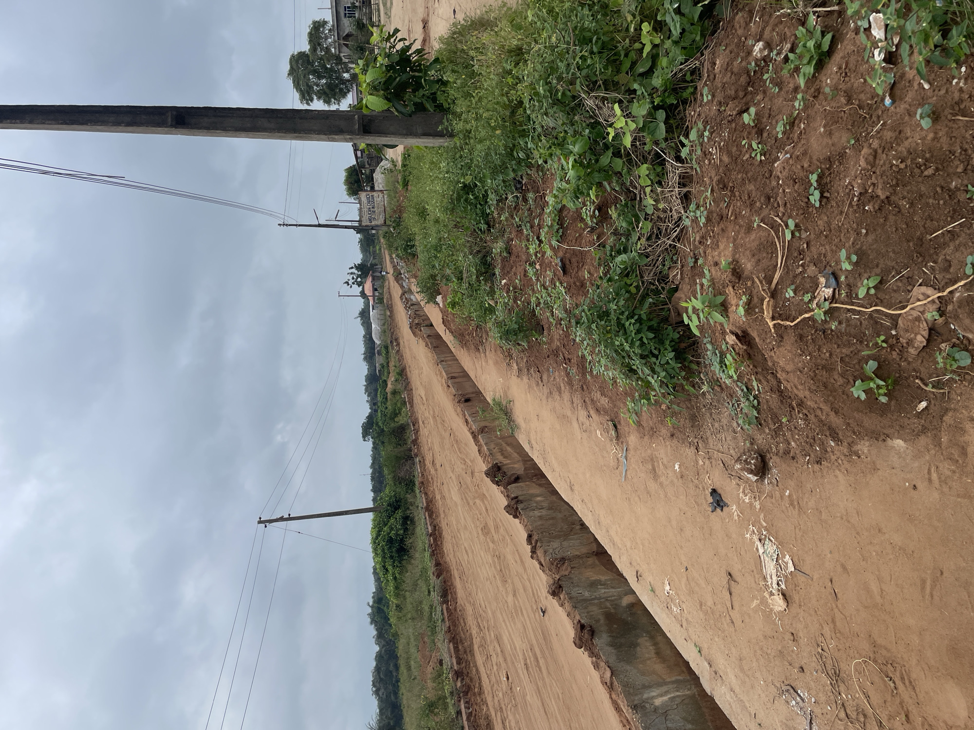 The image depicts a rural scene in Iyafin, Nigeria, on a cloudy day. A dirt road runs diagonally through the frame, with a concrete drainage channel running alongside it. The road appears to be under construction or in disrepair, with loose dirt and debris scattered on its surface. Vegetation, including tall grasses and small trees, lines the sides of the road. In the background, several buildings and power lines are visible, suggesting a sparsely populated area with some infrastructure. There are no people visible in the image, but there is evidence of human activity in the form of discarded trash and construction materials. The overall impression is one of a developing area in a tropical or sub-tropical region.