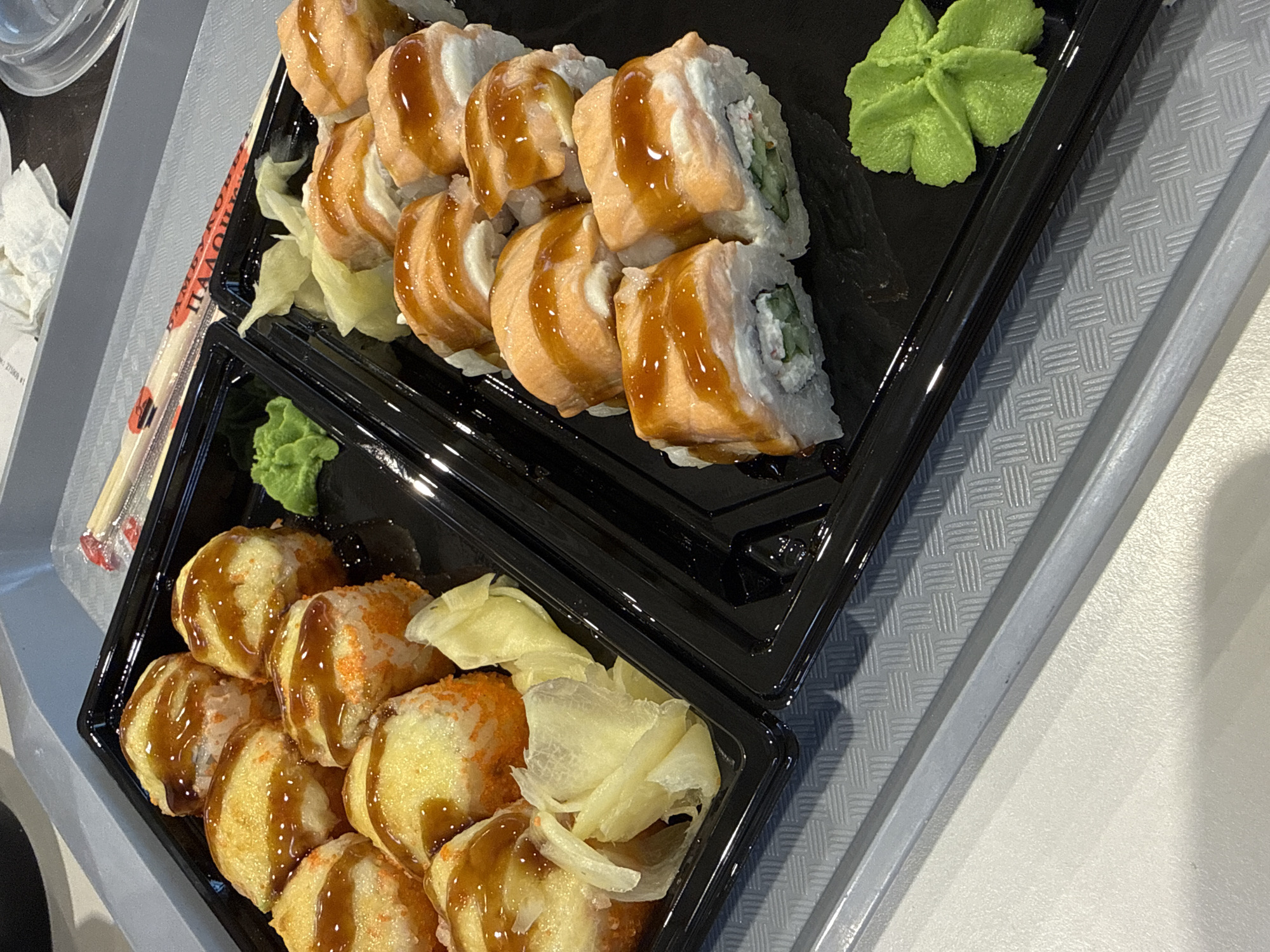 The image shows two black plastic trays of sushi arranged on a gray surface, likely a table or tray. The top tray contains sushi rolls topped with a light orange sauce and drizzled with a brown sauce, alongside pickled ginger and a dollop of bright green wasabi. The bottom tray features different sushi rolls, fried and also drizzled with a brown sauce, garnished with orange roe and pickled ginger. Chopsticks are visible on the side. The sushi likely comes from a restaurant or store in the city of Khimki, Russia, given the location. The scene suggests a meal ready to be consumed.