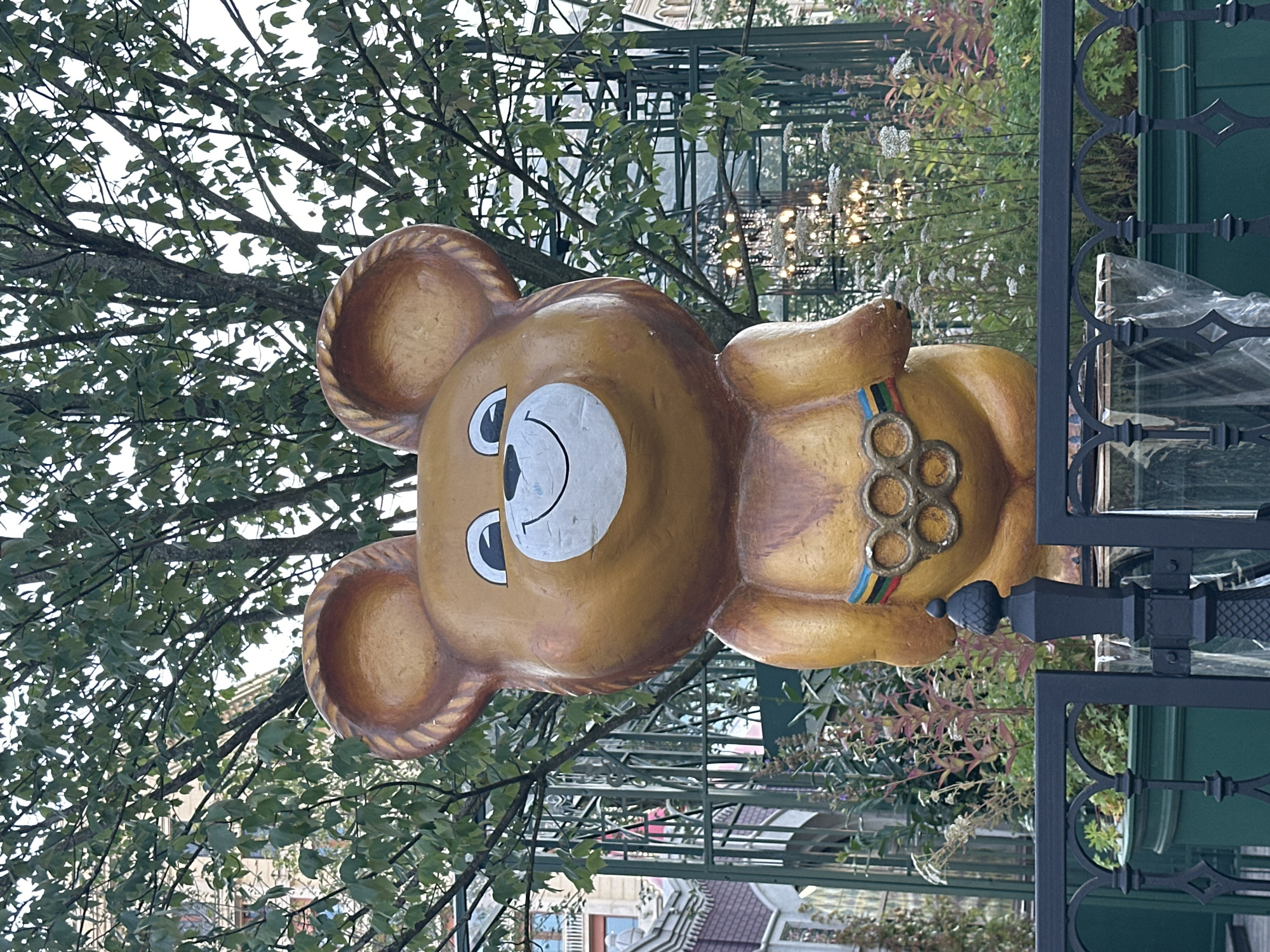 The image shows a large, golden statue of the Olympic mascot "Misha" the bear, from the 1980 Moscow Summer Olympics. The statue is located outdoors, behind a black metal fence. Misha is depicted with a friendly smile and is wearing a belt with the Olympic rings. Behind Misha, there is a lush green tree with visible branches and leaves, and beyond that, a green metal fence. In the background, there are glimpses of buildings and decorative lighting. The overall environment suggests a park or public space in Moscow, Russia, dedicated to commemorating the 1980 Olympic Games.
