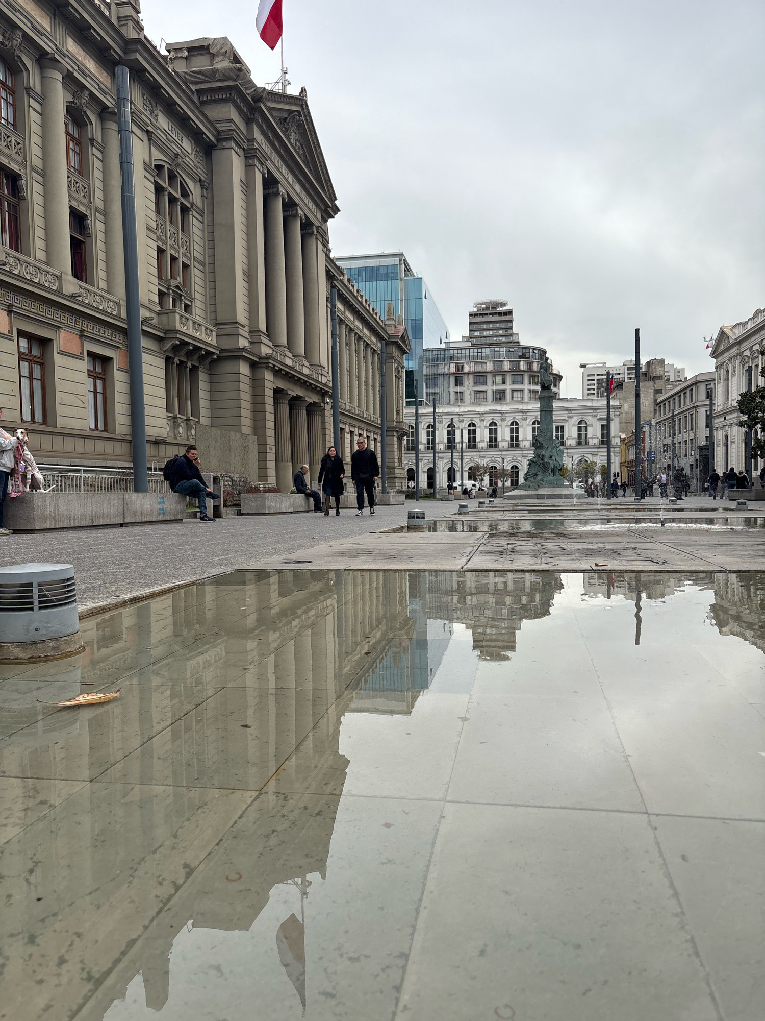 The image captures a wide-angle view of a city square in Santiago, Chile, likely during overcast weather, evidenced by the flat lighting and the presence of puddles reflecting the surrounding architecture. In the foreground, a large puddle of water covers a significant portion of the tiled ground, with reflections of buildings and the sky visible on its surface. A single brown leaf rests on the wet tiles.
To the left, a grand, old building with classical architectural elements dominates the scene. It features a stone facade, tall columns, arched windows with dark frames, and a triangular pediment. A flag with red and white stripes, possibly the Chilean flag, is visible flying from a pole on top of this building. A dark gray lamppost stands in front of it.
Further into the square, more modern buildings are visible, including a glass-fronted building. In the mid-ground, a prominent monument or statue stands atop a tall column. Several people are scattered throughout the square, some walking, some sitting on a low concrete wall. Among them are two individuals walking together, a person seated on a bench talking on a phone, and a group of people with two dogs near the building on the left. The ground is paved with tiles and cobblestones. In the background, the cityscape extends with more buildings of varying architectural styles. The overall impression is that of a public space in a historical city center.