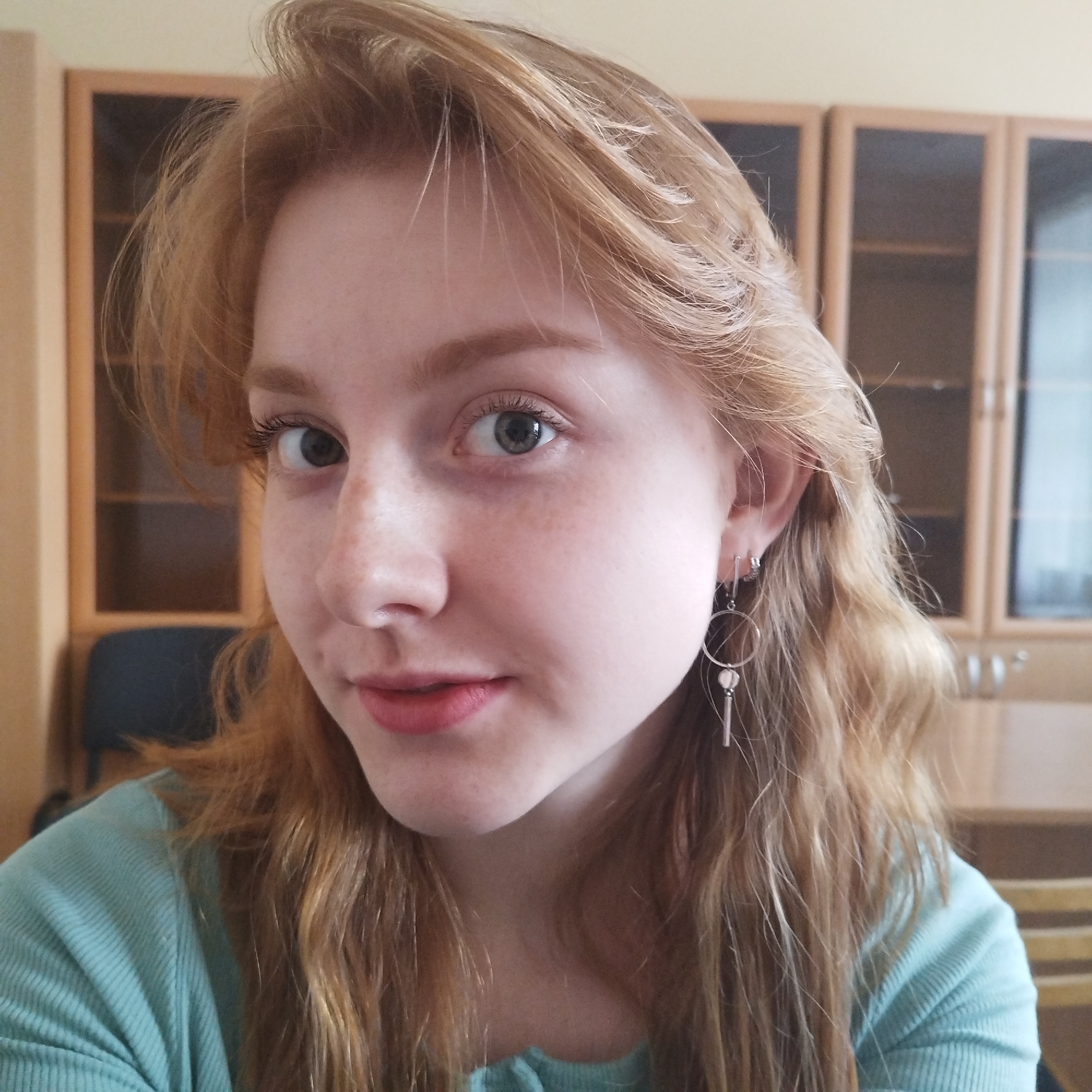 A young woman with light red, wavy hair and fair skin, adorned with freckles, is captured in a close-up portrait. Her eyes, appearing a greenish-grey hue, look directly at the viewer. She has a soft, slight smile on her lips, which are painted a subtle reddish-pink. She wears a light blue or teal ribbed top. On her right ear, she has two silver earrings: one is a simple small hoop, and the other is a larger hoop with a small circular charm and a dangling bar beneath it.
The setting appears to be indoors, with a warm-toned, blurred background. Behind her, out of focus, are wooden cabinets with glass doors, suggesting an office, study, or classroom environment. The lighting seems natural and soft, indicating daytime. There is no discernible text in the image. The overall mood conveyed is calm and approachable.