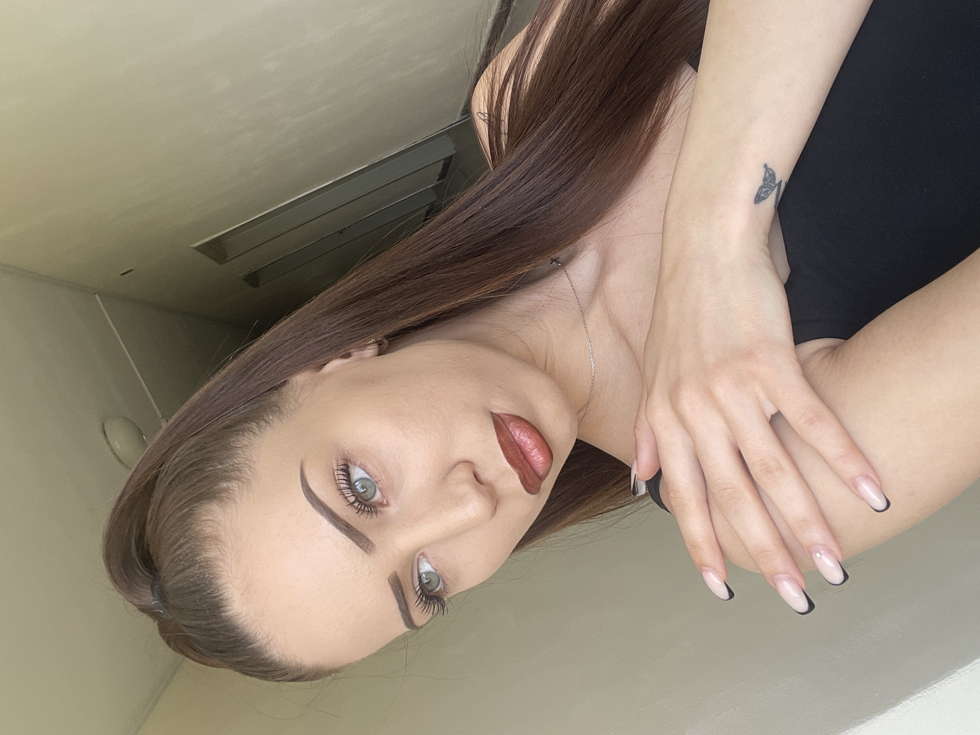 This is an indoor portrait of a young woman, taken in Veliko Tarnovo, Bulgaria. She is positioned on her side, looking directly at the camera with a neutral expression. She has long, straight dark brown hair that falls over her shoulder, light-colored (possibly blue-green) eyes, well-defined dark eyebrows, dark mascara, and reddish-brown lipstick.
She is wearing a dark, possibly black, top and a thin silver chain necklace. Her arms are crossed, with her left hand resting on her right arm. Her nails are long and manicured in a French tip style with black tips. A small, black butterfly tattoo is visible on her right forearm/wrist.
The background consists of light-colored walls and a ceiling, with horizontal recessed light fixtures visible in the upper right background, suggesting a modern interior space. The lighting is even, illuminating the subject's face clearly. There is no visible text in the image. The setting appears to be indoors during what could be daytime or well-lit artificial conditions.