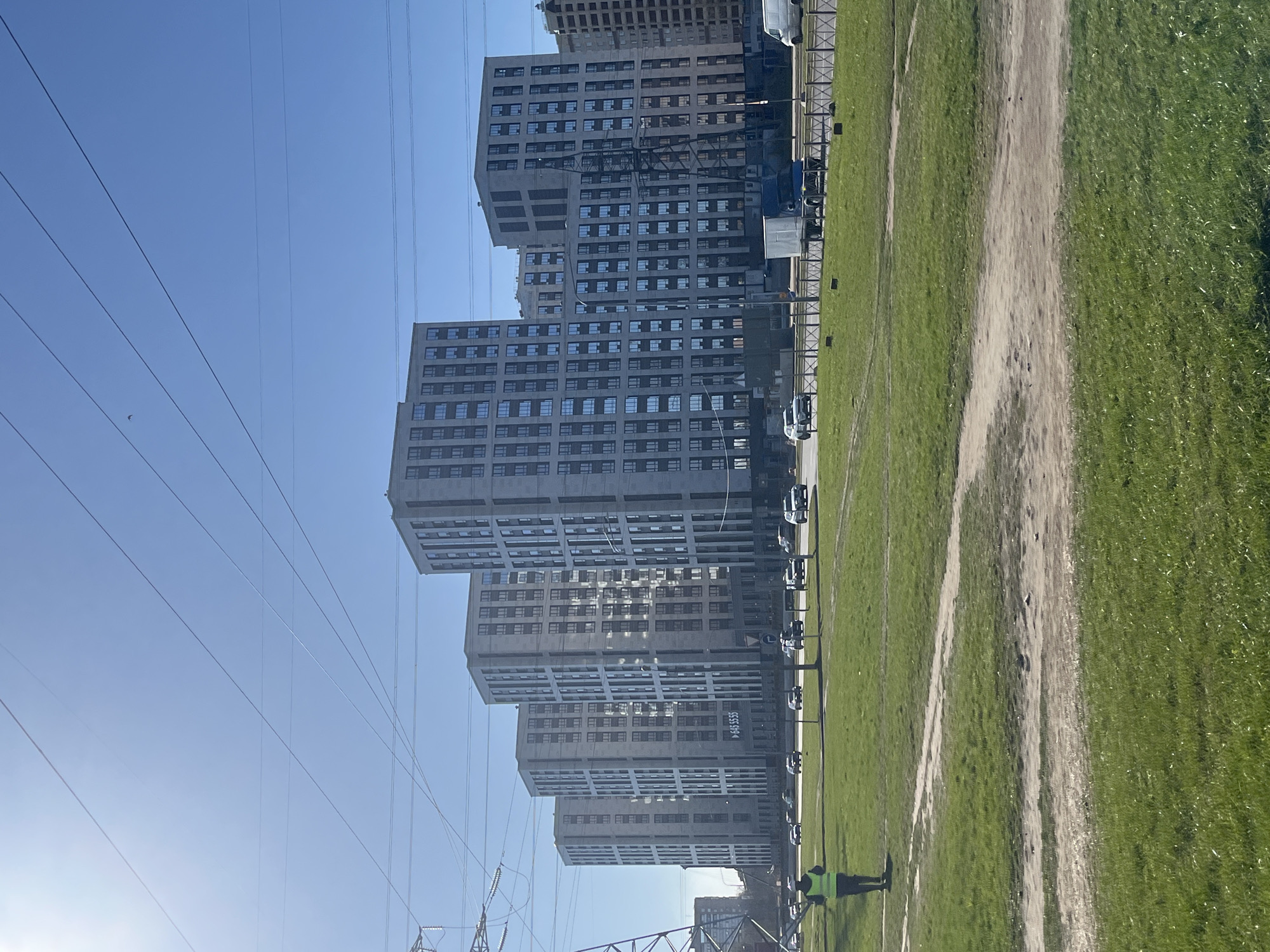 The image shows a row of modern apartment buildings in Saint Petersburg, Russia. The buildings are tall, with a repetitive pattern of windows. A grassy field with dirt paths borders the buildings. A row of parked vans is visible along the street. A person wearing a high-visibility vest stands near the edge of the field. Power lines and pylons are visible in the foreground against a clear blue sky.