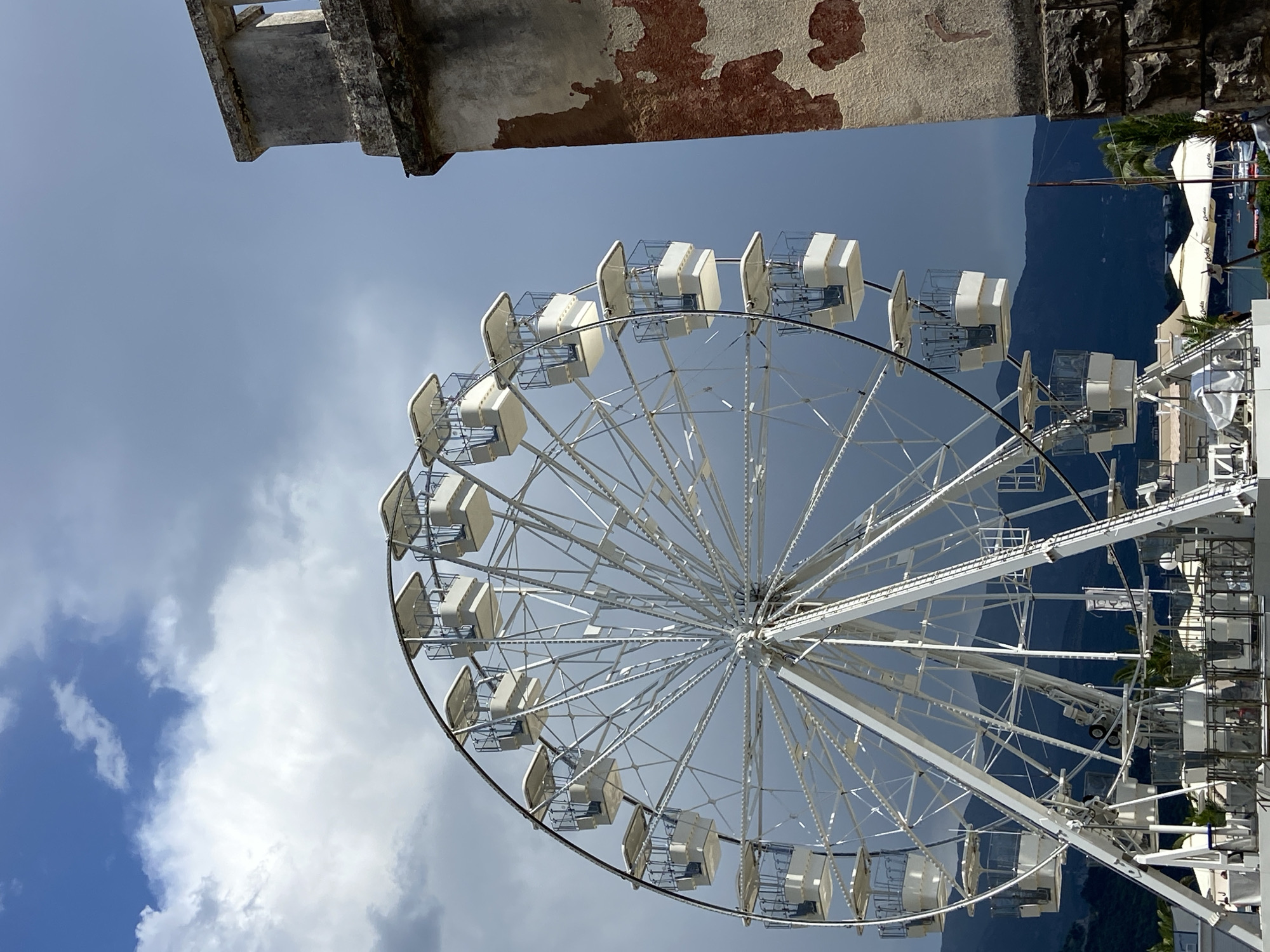 The media file shows a large white Ferris wheel against a cloudy sky with dark mountains in the background. The Ferris wheel is the primary subject, with its intricate metal structure and numerous white cabins visible. Some cabins have clear plastic enclosures.
In the upper left corner, a portion of an old stone structure with peeling paint is visible, suggesting a historical or weathered element in the environment. The sky is a mix of blue and gray clouds, indicating potentially variable weather.
The image is taken from a low angle, looking up at the Ferris wheel. The mountains in the background are dark and imposing, possibly suggesting a coastal or mountainous region. Some greenery, possibly palm trees, and white umbrellas are visible at the base of the Ferris wheel on the right side, hinting at a resort or leisure area.
There is no clear indication of people or a specific event taking place. The time of day appears to be daytime, given the brightness of the sky. The overall mood is somewhat dramatic due to the contrast between the bright Ferris wheel and the dark sky and mountains.
No visible text is present in the image. The location context provided is Budva, Montenegro, which aligns with the possibility of a coastal resort with historical architecture and mountainous surroundings.