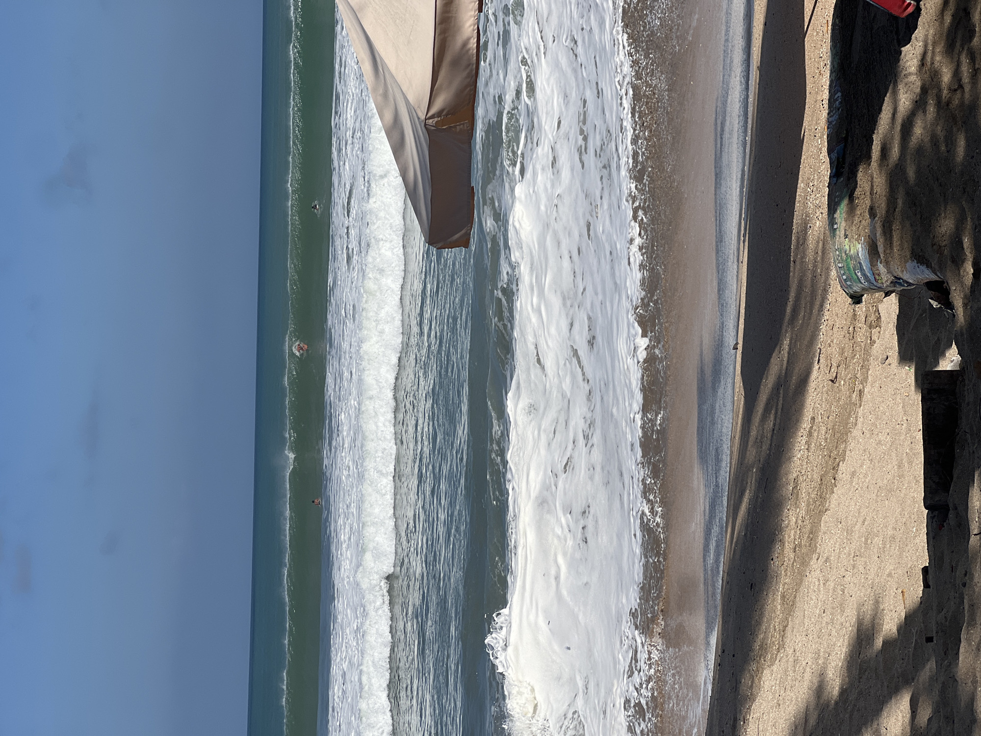 The image captures a view of the ocean at Kuta Beach, Bali, Indonesia. The foreground shows a sandy beach with shadows cast by trees, and some debris is visible on the sand. In the middle ground, foamy white waves crash onto the shore, with the dark sand of the beach appearing wet from the tide. Further out, calmer, darker blue water is visible, interspersed with several lines of white-capped waves. In the distance, several people are in the water, with at least two appearing to be surfing. The sky is a clear, bright blue with a few faint clouds. A portion of a large, tan beach umbrella is visible on the right side of the frame, providing shade to the beach area. The overall scene depicts a sunny day at a popular surfing beach.