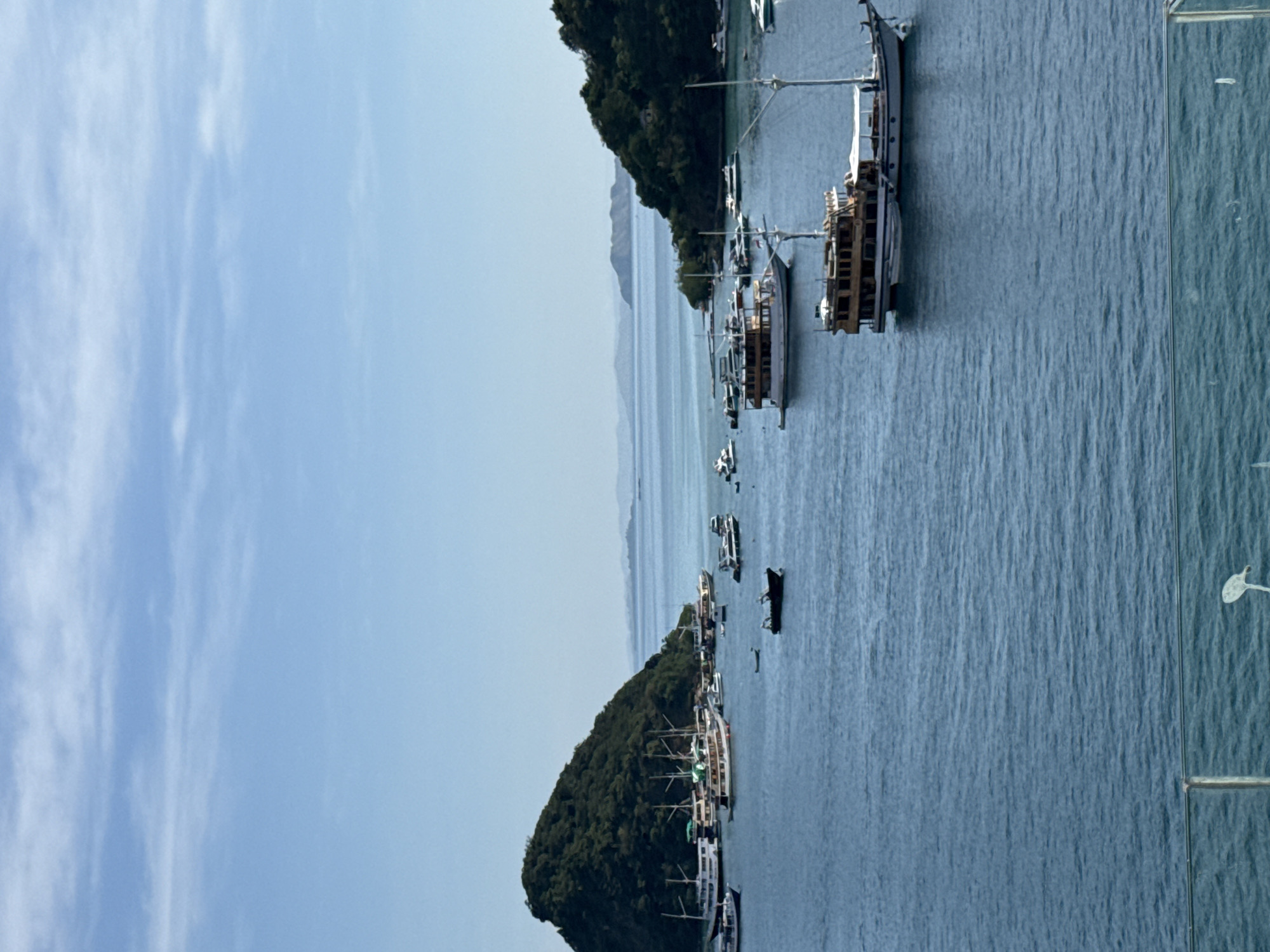 The image shows a view of the sea in Labuan Bajo, Indonesia. There are several boats anchored in the water near a small island covered in lush green vegetation. The sky is blue with some scattered clouds. In the distance, there are more islands and a hazy horizon. The photograph is taken from an elevated position, likely a hotel or building, and part of a railing or structure is visible in the right side.