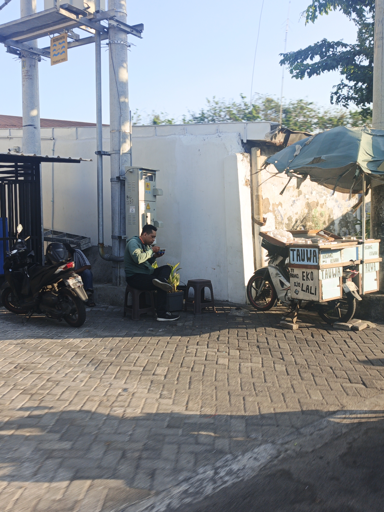 The image depicts a street scene in Surabaya, Indonesia. In the foreground, a man is seated on a stool, eating from a bowl. He is wearing a green hoodie and black pants. A plant in a pot sits next to him. To his left, a black motorcycle is parked. Further to the left, another motorcycle is visible. Behind the man, a white wall serves as a backdrop, with a utility box mounted on a pole. To the right of the man, a food cart attached to a motorcycle is parked under a damaged awning. The cart has various labels, including "TAUWA," "EKO," and "LALI." The ground is paved with interlocking bricks, and shadows are cast by the objects and trees. In the background, trees and a clear blue sky are visible. A yellow warning sign with Indonesian text and wavy symbols is attached to a utility pole on the left side of the image.