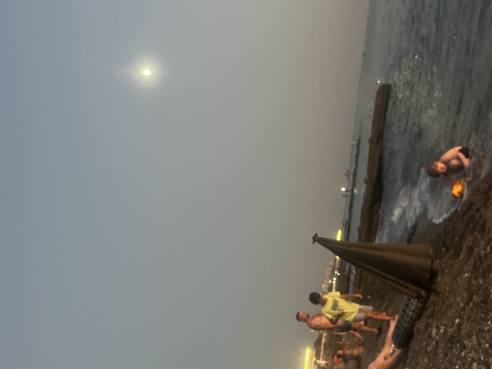 This media file depicts a twilight scene on a pebble beach in Sochi, Russia. The sky is a soft grey, with a bright, hazy moon visible in the upper left quadrant. The water is dark and calm, with small waves lapping at the shore.
In the foreground, several people are visible. Two young men, one wearing a yellow t-shirt and the other shirtless, stand near a dark, cone-shaped object that appears to be a beach umbrella or shade structure. Another person, also shirtless, is partially visible behind them, along with another figure whose legs are showing. Further down the beach, a child is playing in the shallow water. They are mostly submerged, with only their head and shoulders visible, and appear to be interacting with something in the water.
The overall atmosphere is relaxed and casual, suggesting a late evening spent at the beach. The lighting indicates that it is dusk or early evening. Distant lights of buildings can be seen along the coastline in the background, providing a hint of urban proximity within the natural setting of the beach. No visible text is present in the media file.